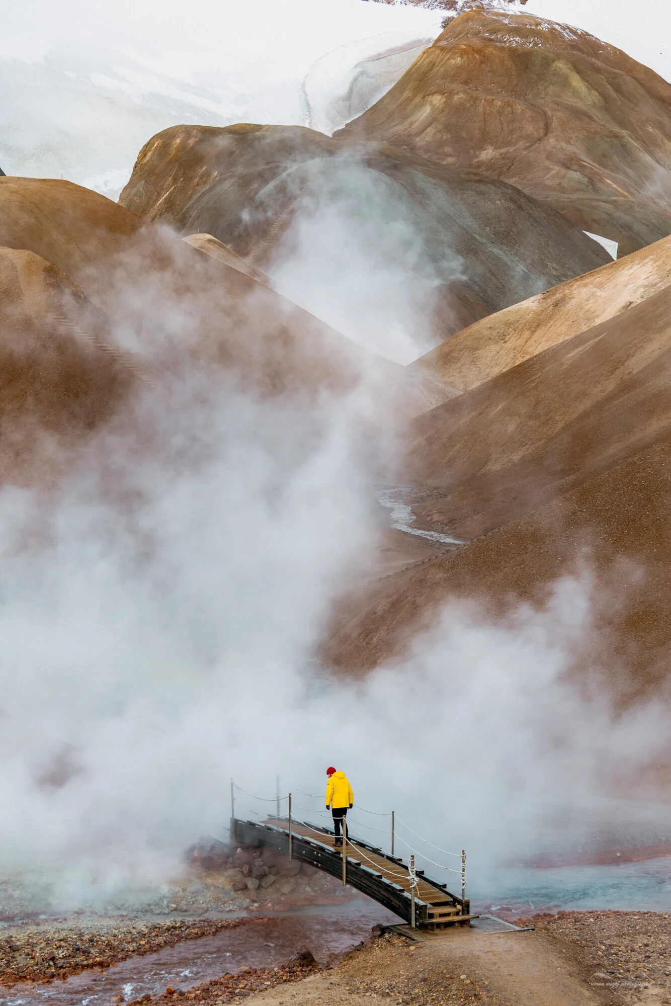 A person in a yellow jacket and red hat walking on a wooden bridge in a geothermal area with steaming, colorful volcanic hills in the background.