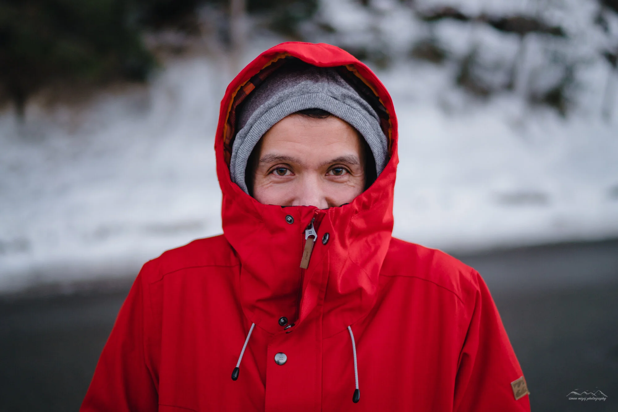 A person wearing a red winter jacket with the hood up, a gray beanie underneath, smiling in an outdoor winter setting with snow and blurred trees in the background.