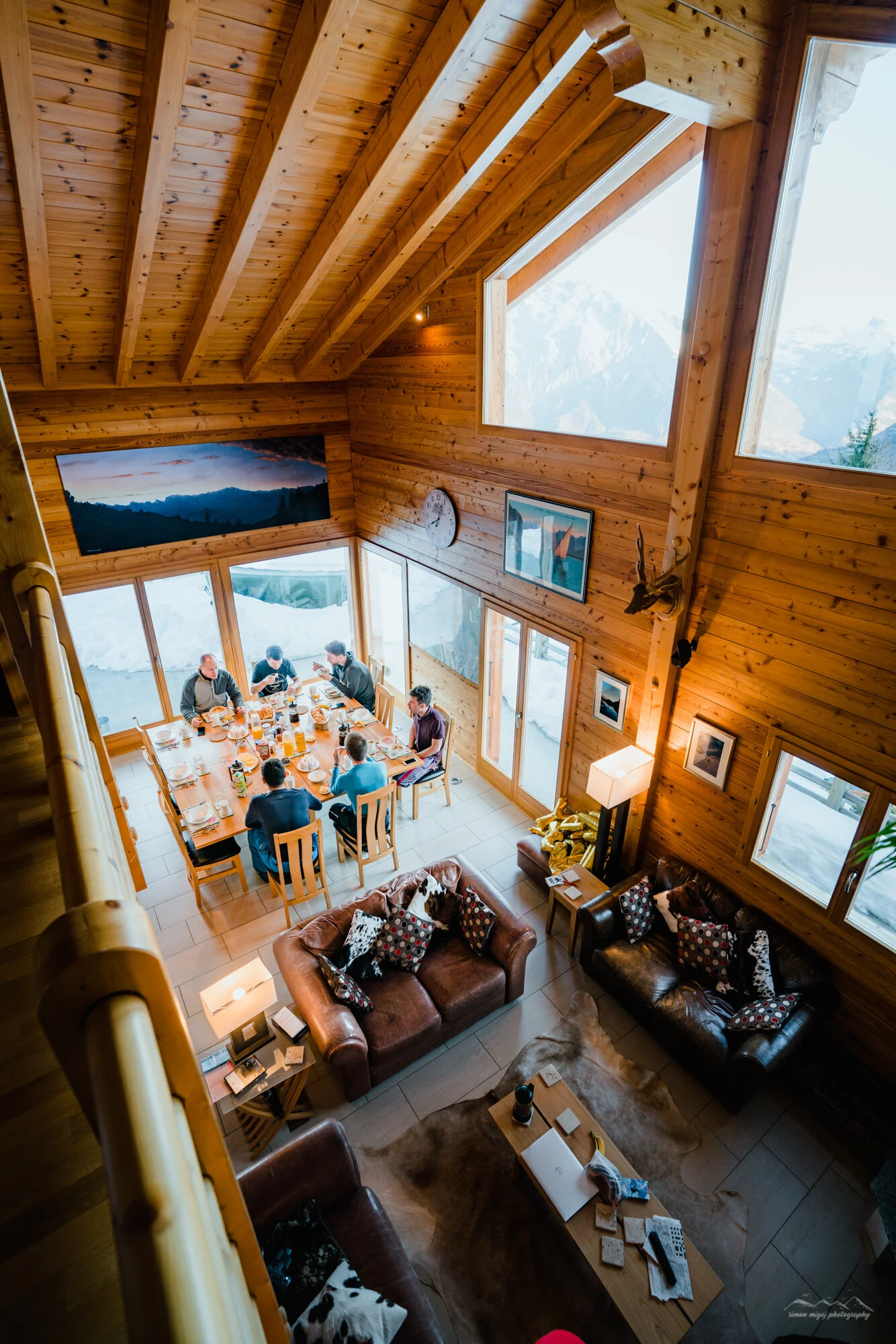 A cozy mountain cabin interior with wooden walls and ceiling, large windows showing snowy mountain scenery, a group of people sitting at a dining table set for a meal, and a living area with leather sofas decorated with patterned cushions.