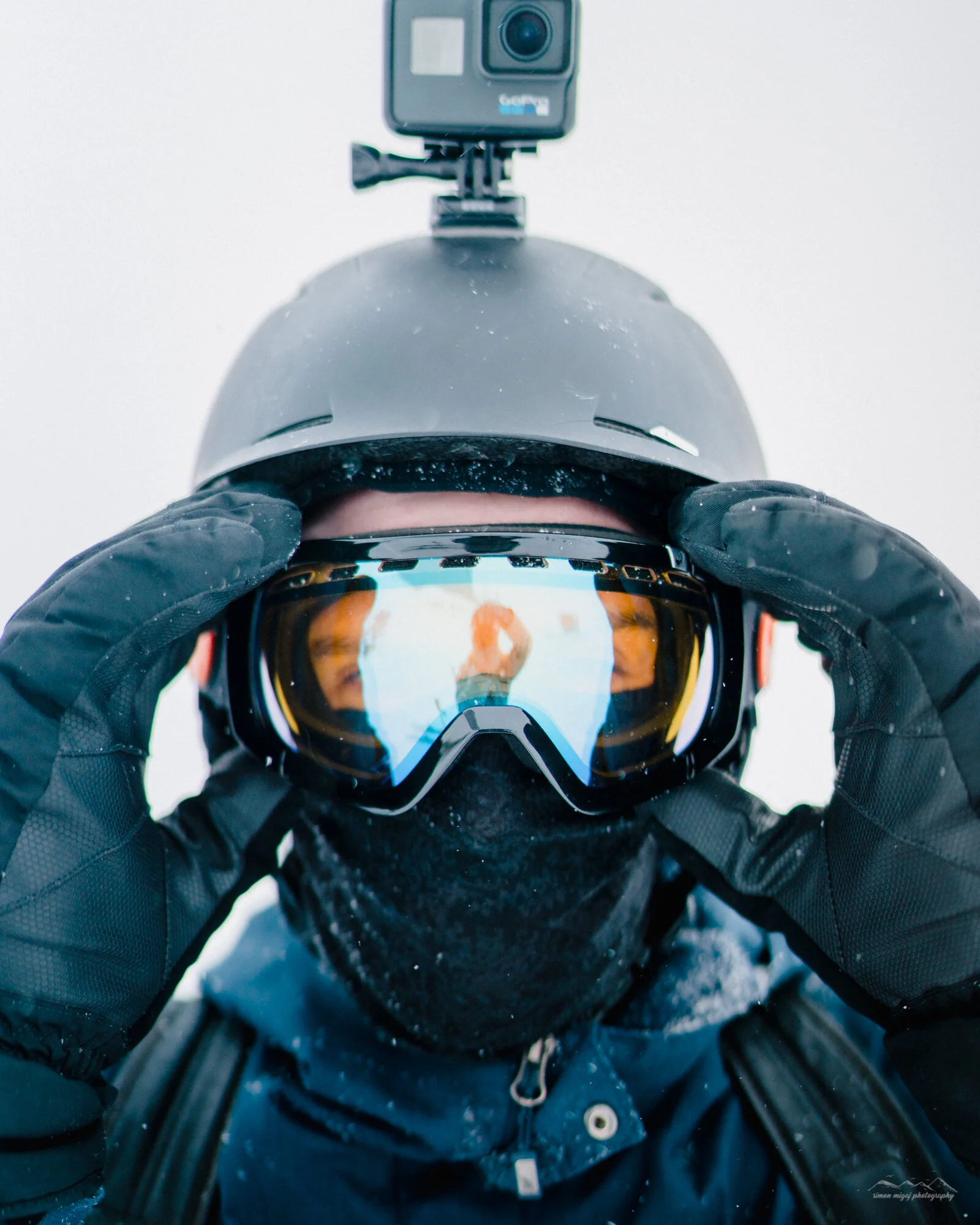 Person wearing a helmet and ski goggles, taking a selfie in a snowy environment, with snow on the helmet and wearing black gloves and winter gear.