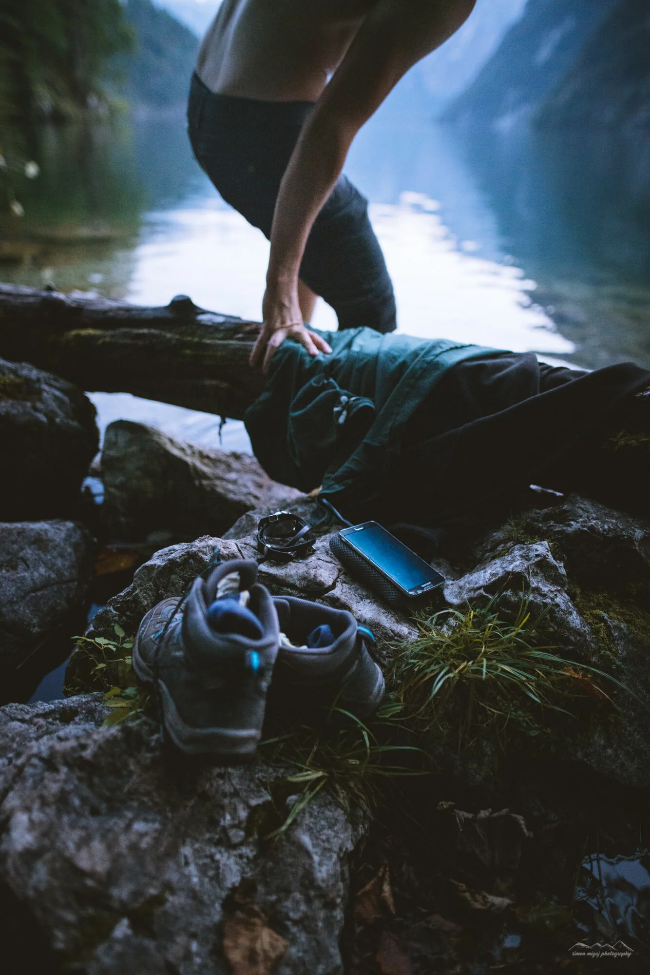 A person in black shorts and a tank top is bending over on a large fallen log by a river, with mountains and trees in the background. There are hiking shoes, a smartphone, and outdoor gear on the rocks near the water.
