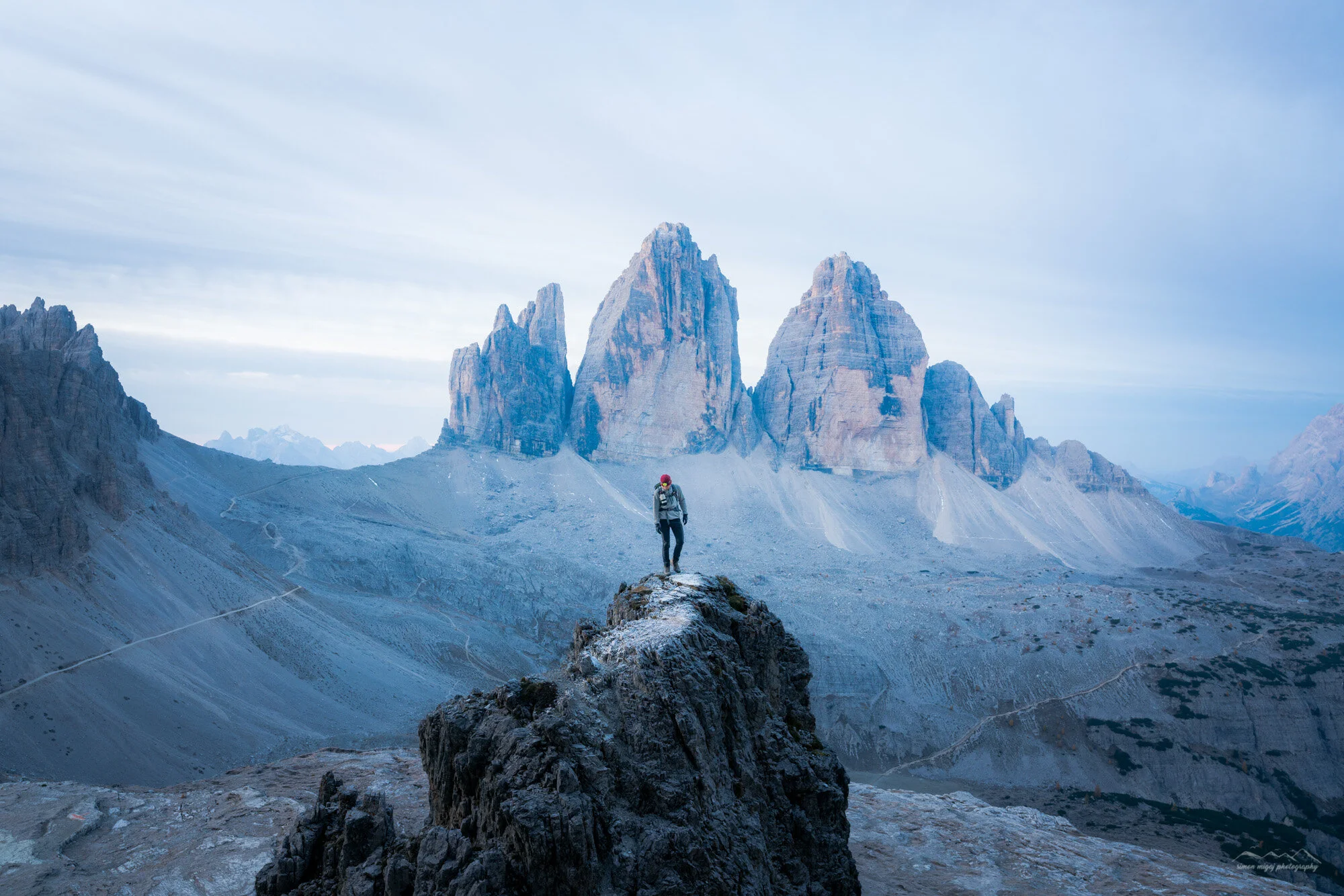 A lone hiker in a red helmet and gray jacket walking across a rocky, mountainous landscape with towering jagged peaks in the background under an overcast sky.
