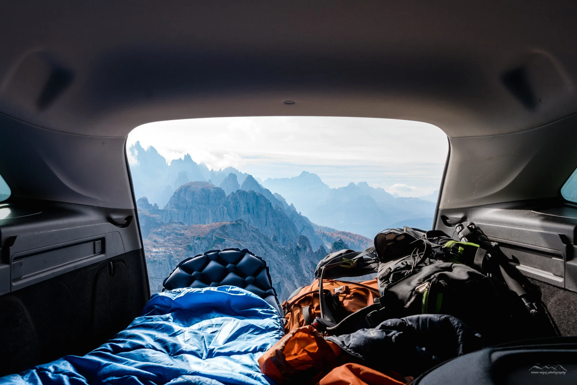 View from inside a vehicle trunk showing mountain landscape through open hatch, with scattered camping gear and clothing.