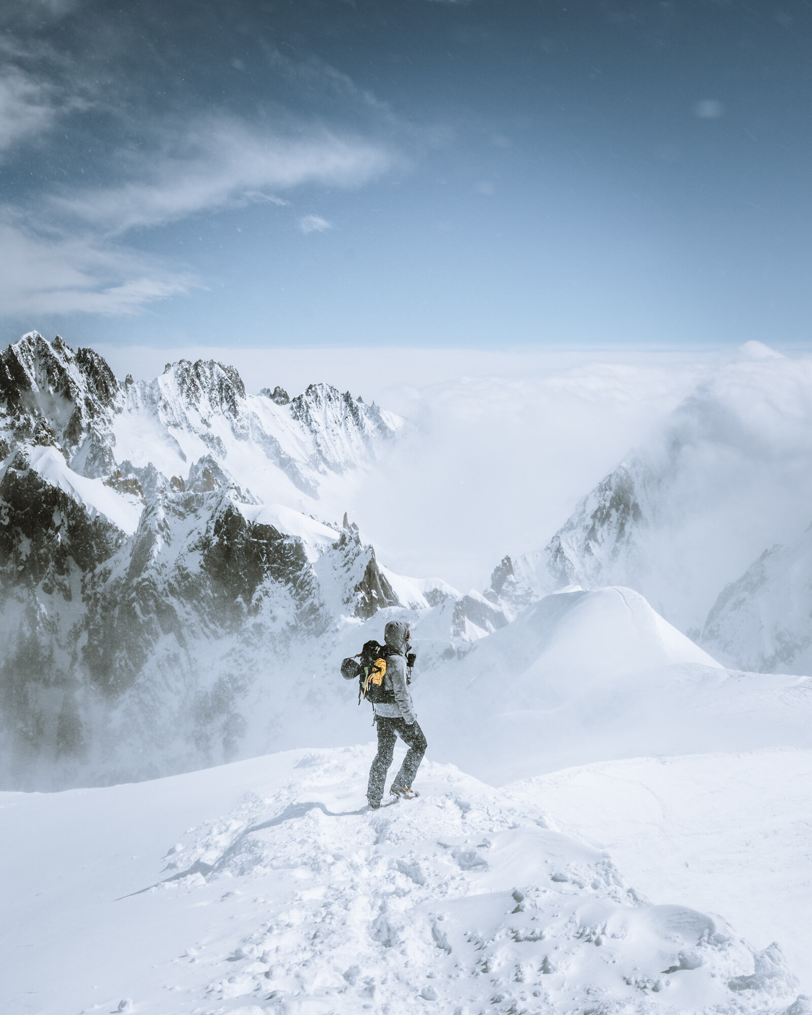 A person with a backpack stands in a snowy mountain landscape, with jagged snow-covered peaks and a cloudy sky in the background.