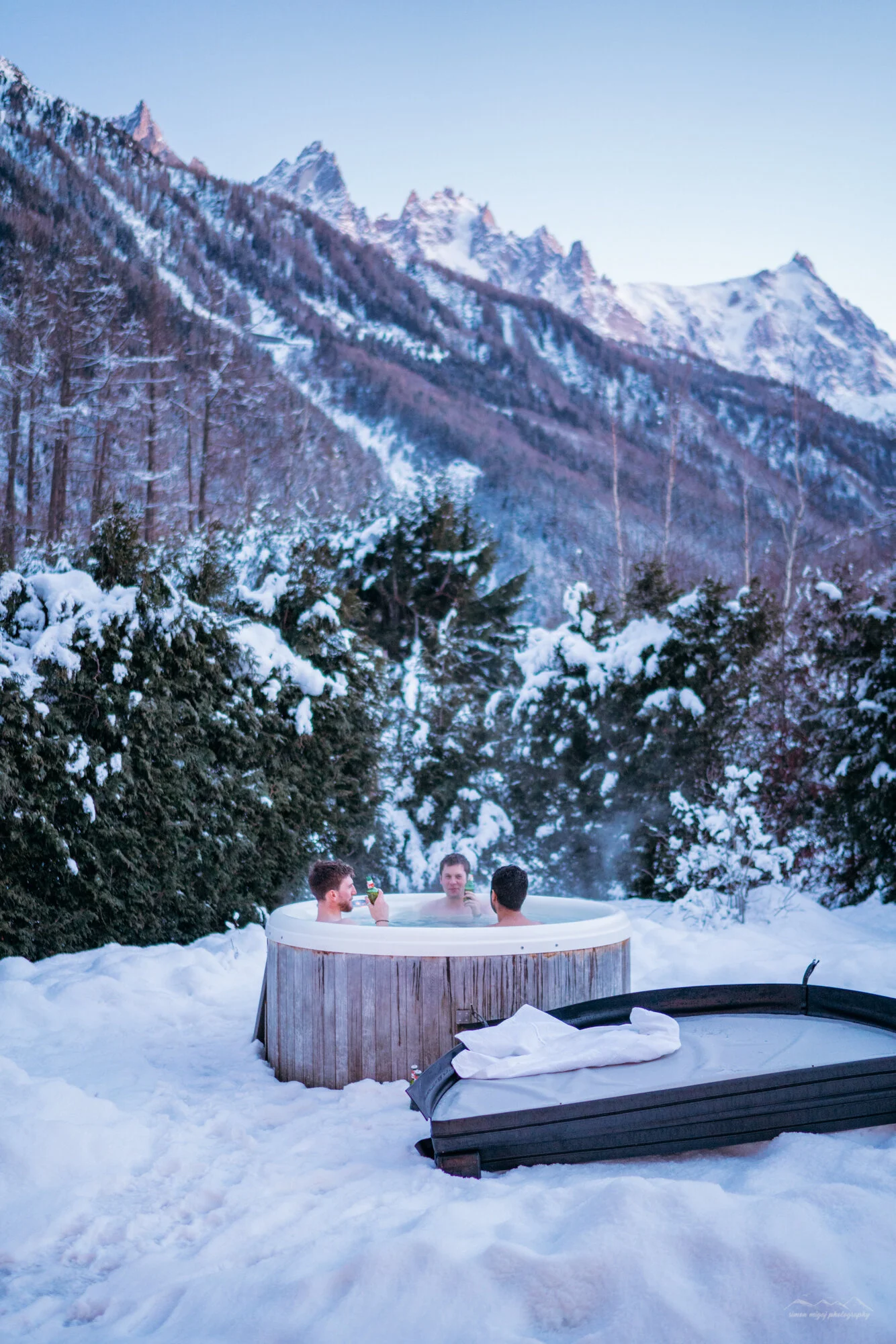 Four men sitting in a hot tub outdoors in a snowy landscape, surrounded by snow-covered trees and mountainous terrain in the background, with one man holding a drink.