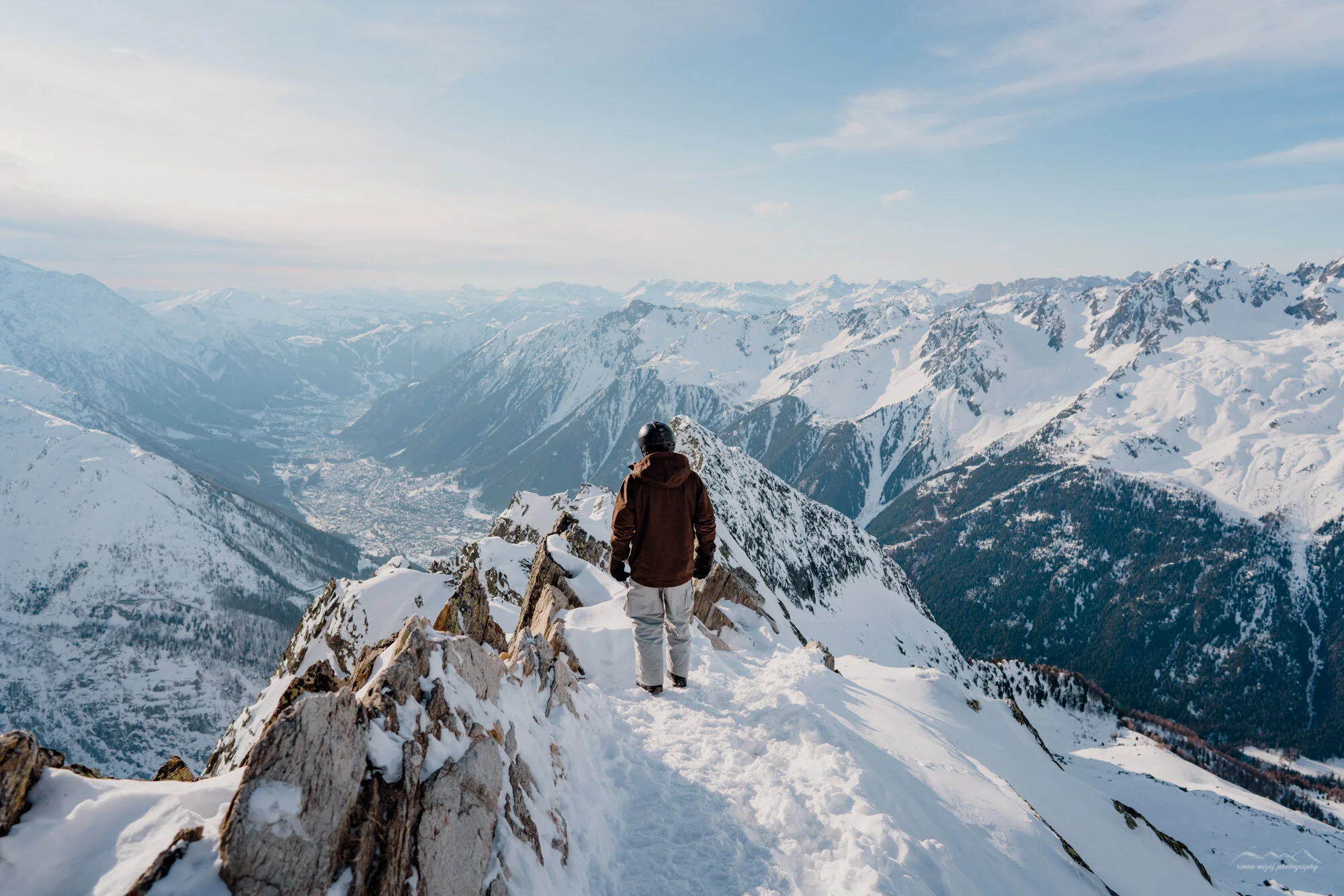 A person wearing a brown jacket, light gray pants, and a black helmet stands on snow-covered rocks at a mountain summit, looking out over snow-capped mountain ranges and a valley below under a partly cloudy sky.