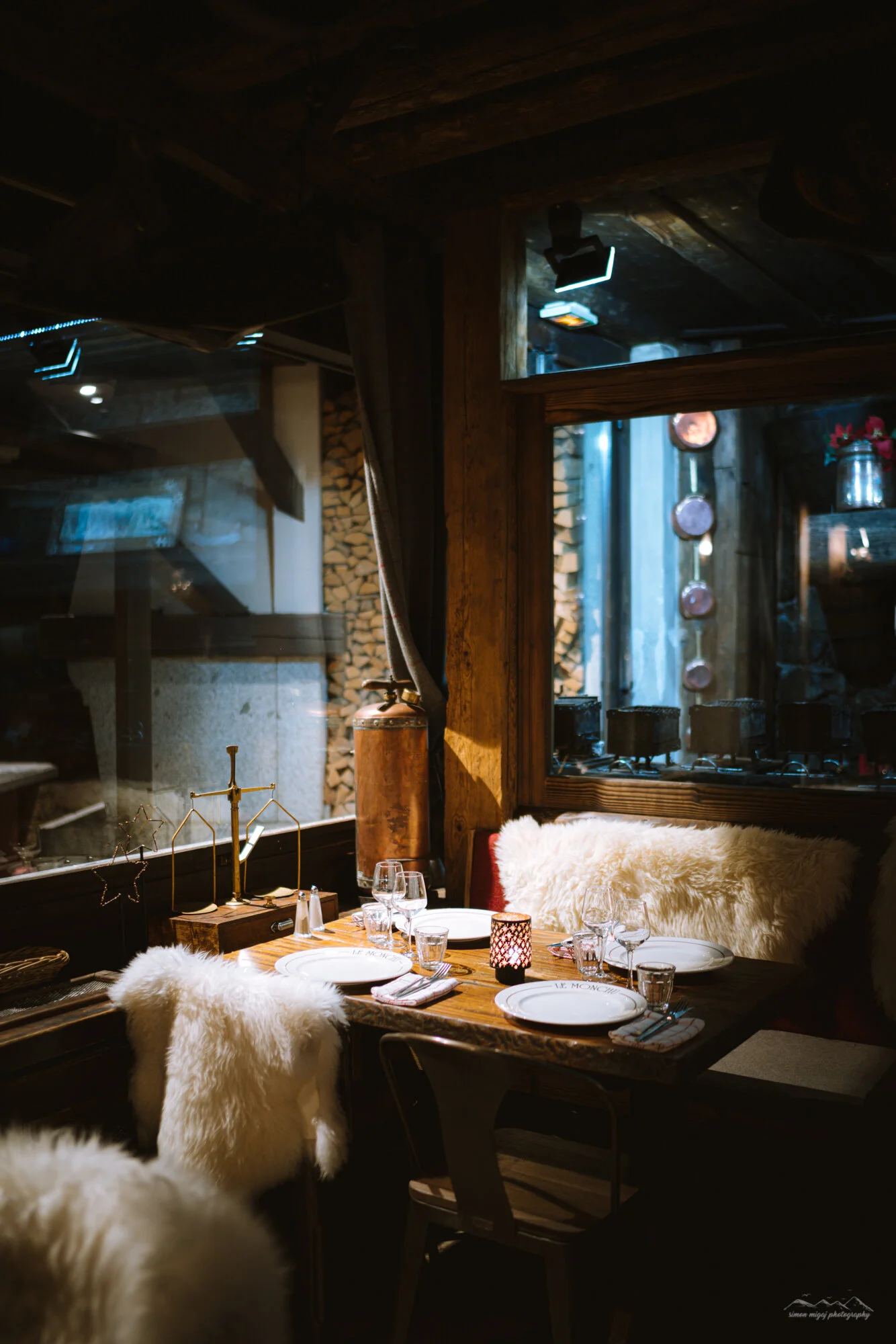 A cozy rustic dining room featuring a wooden table set for a meal with white plates, wine glasses, and a black candle holder with a lit candle. The table has plush faux fur chair cushions and decorative star-shaped ornaments. Large windows reveal a w