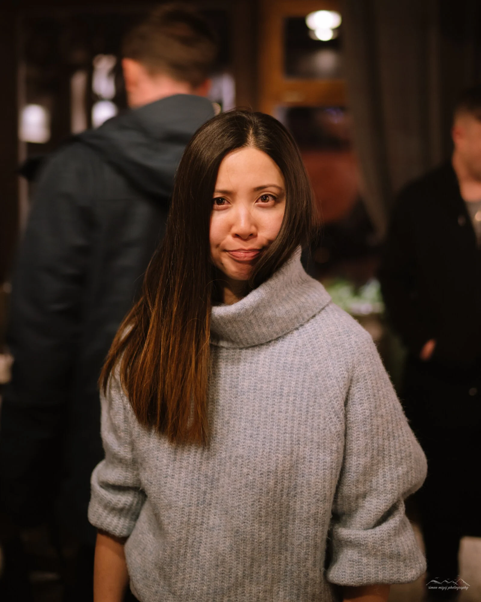 A woman with long brown hair wearing a gray knitted sweater, standing indoors with blurred background of people.