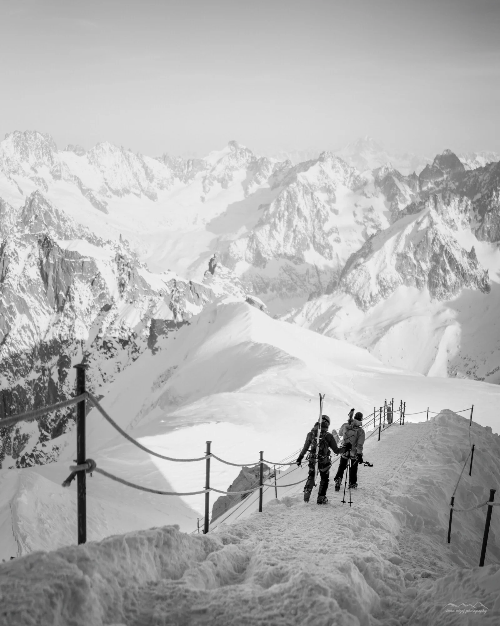 Snow-covered mountain landscape with two skiers walking along a trail and a safety fence in the foreground.