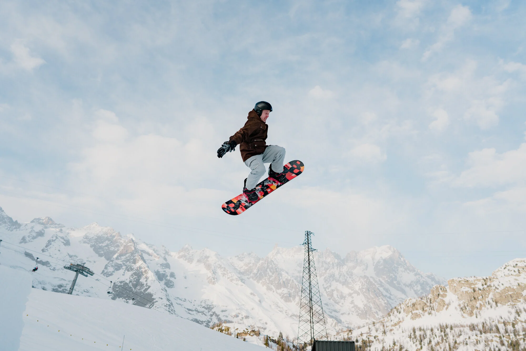 A person wearing a helmet and winter jacket snowboarding in the air against a backdrop of snow-covered mountains and cloudy sky, with ski lifts and towers in the background.