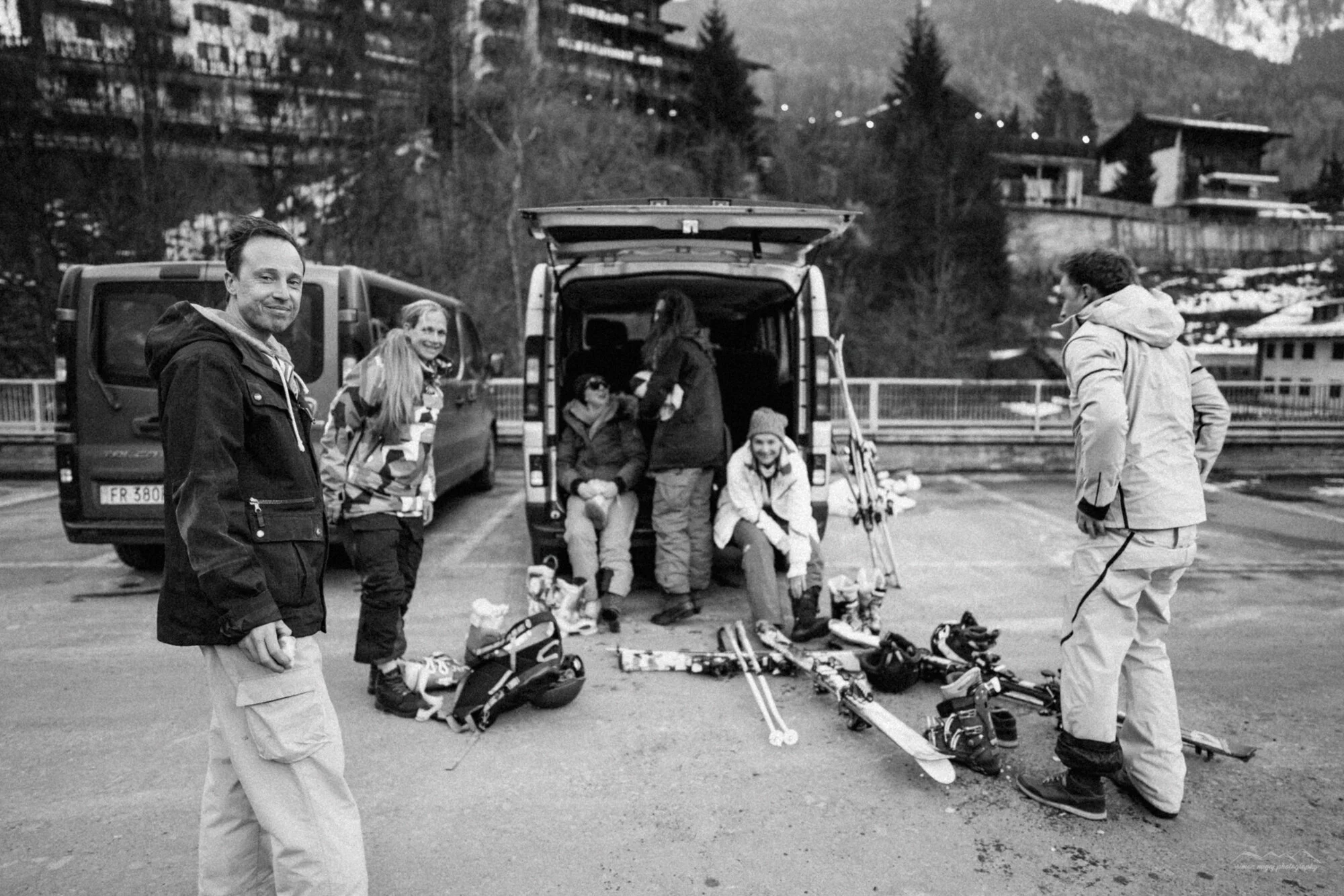 A group of people preparing for skiing outdoors near a parking lot with mountains in the background. They are surrounded by ski gear and equipment.