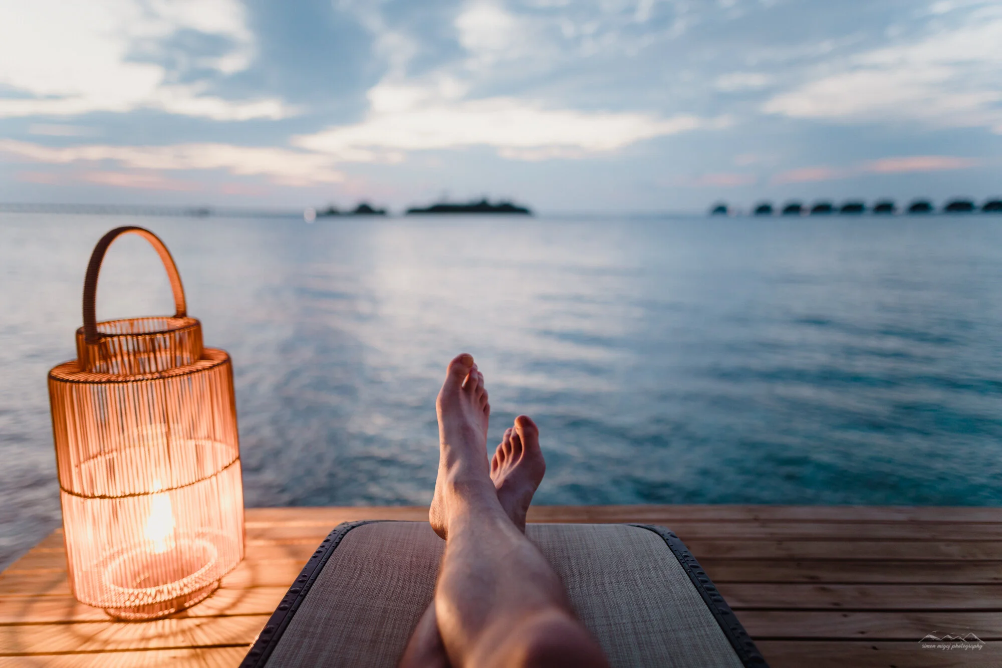 Person relaxing on a lounge chair by the water during sunset with a lantern nearby and a blurry island in the distance.
