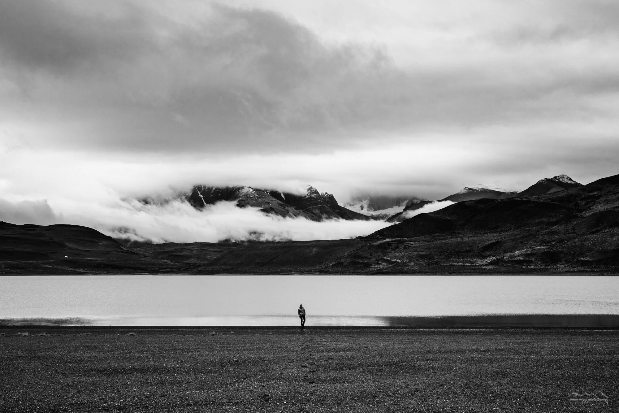 A person standing alone on a flat, open landscape with large mountains and clouds in the background, in black and white.