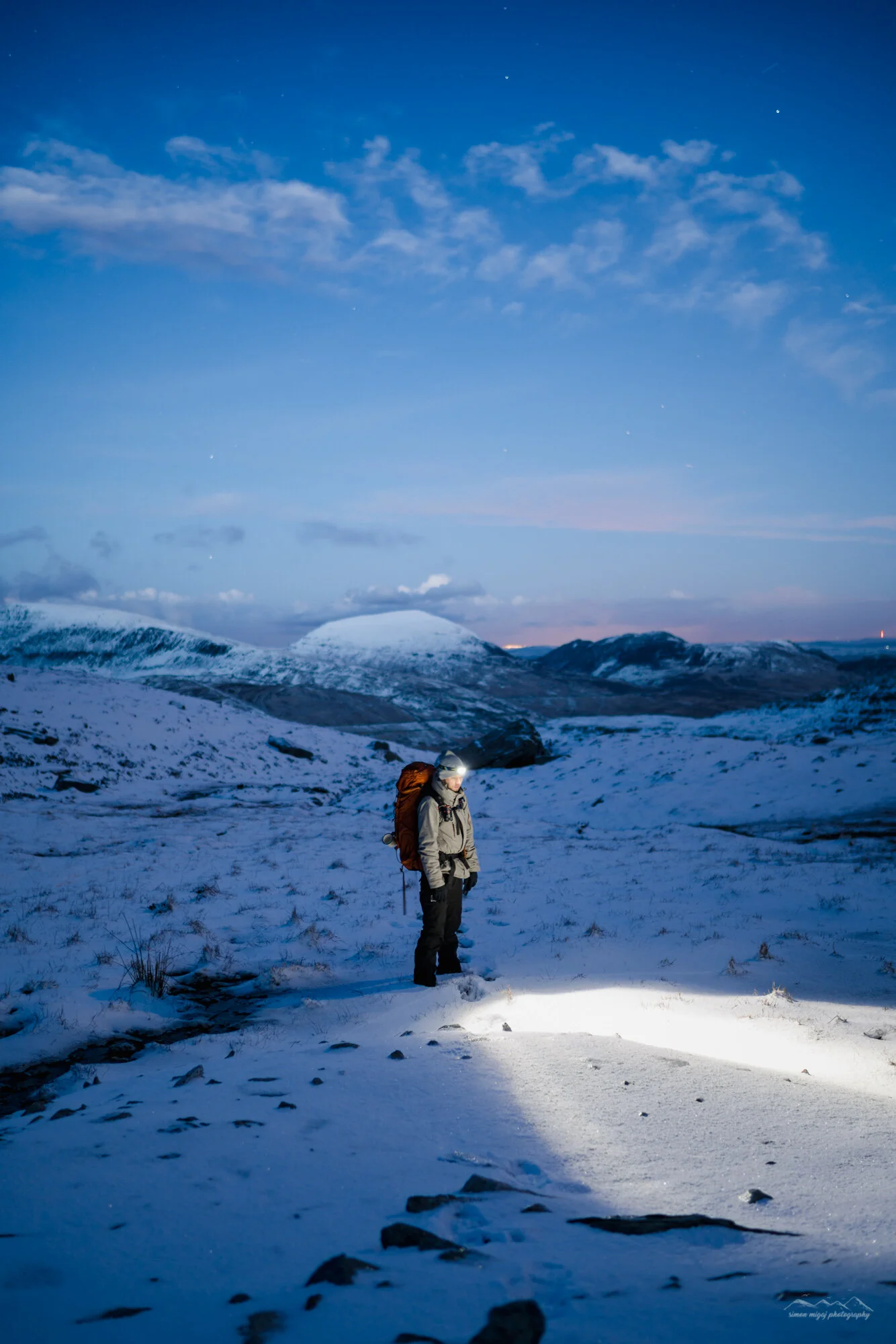 A lone hiker dressed in winter gear, including a gray jacket, black pants, and a headlamp, standing in a snowy mountainous landscape during twilight with snow-covered mountains in the background and a darkening sky with some clouds.