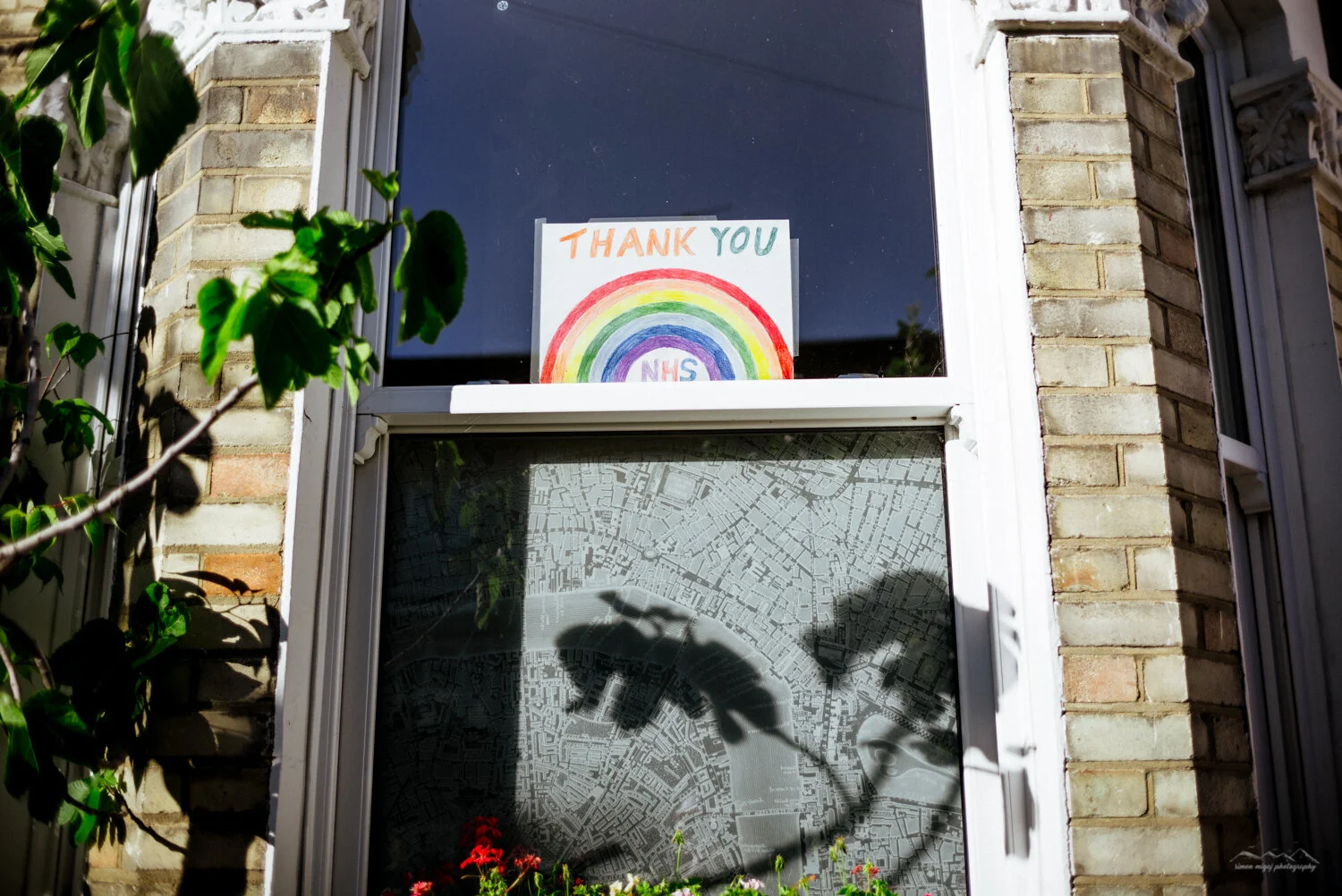 A windows with a handmade sign that says 'Thank You NHS' and features a rainbow drawing. The window also shows a map and shadows of a plant and a person's hand holding a phone outside.