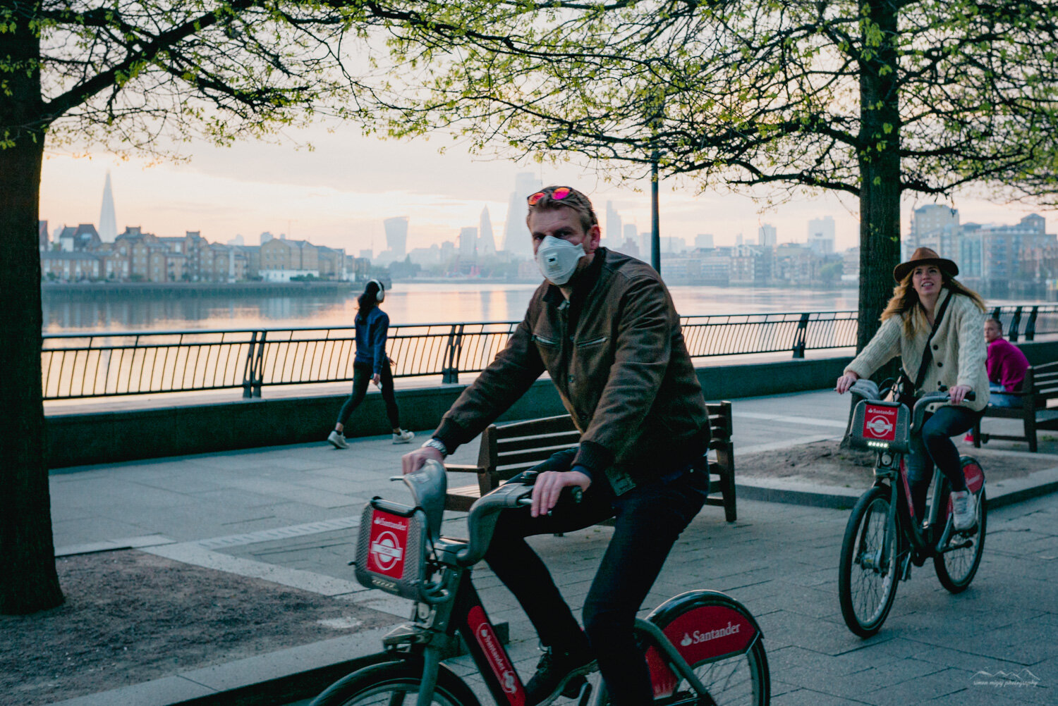 People cycling along a riverside promenade in an urban area with a city skyline in the background. The man in the foreground is wearing a mask, jacket, and sunglasses, while the woman behind him is wearing a hat and smiling.