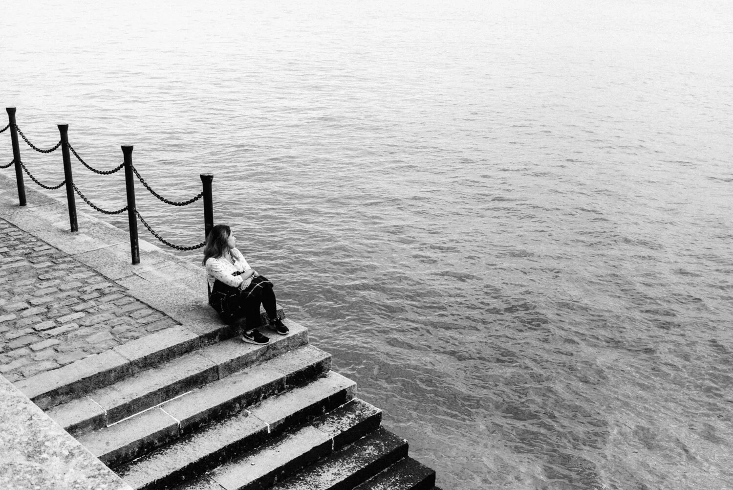 A young woman sitting on concrete steps by a body of water, looking out at the water, in black and white.