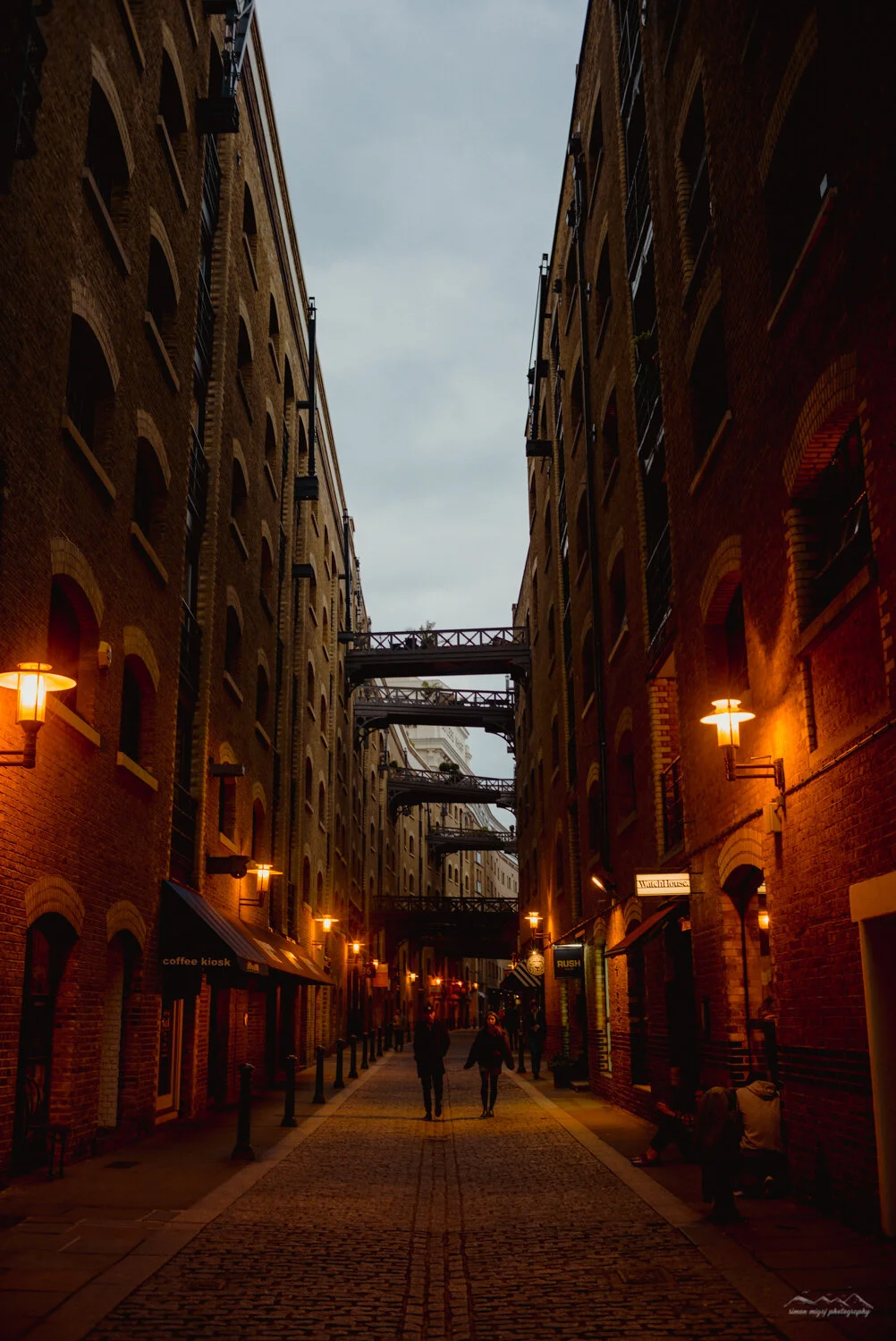 A narrow street between brick buildings with lit street lamps and a few people walking at dusk or early evening.