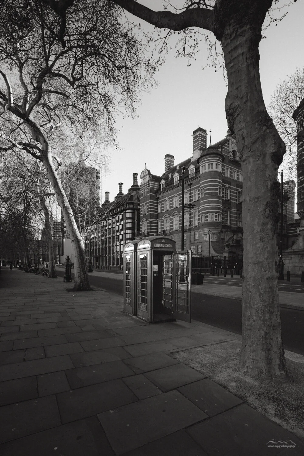 Black and white photo of a street with a traditional British telephone booth, leafless trees, and historic buildings in the background.