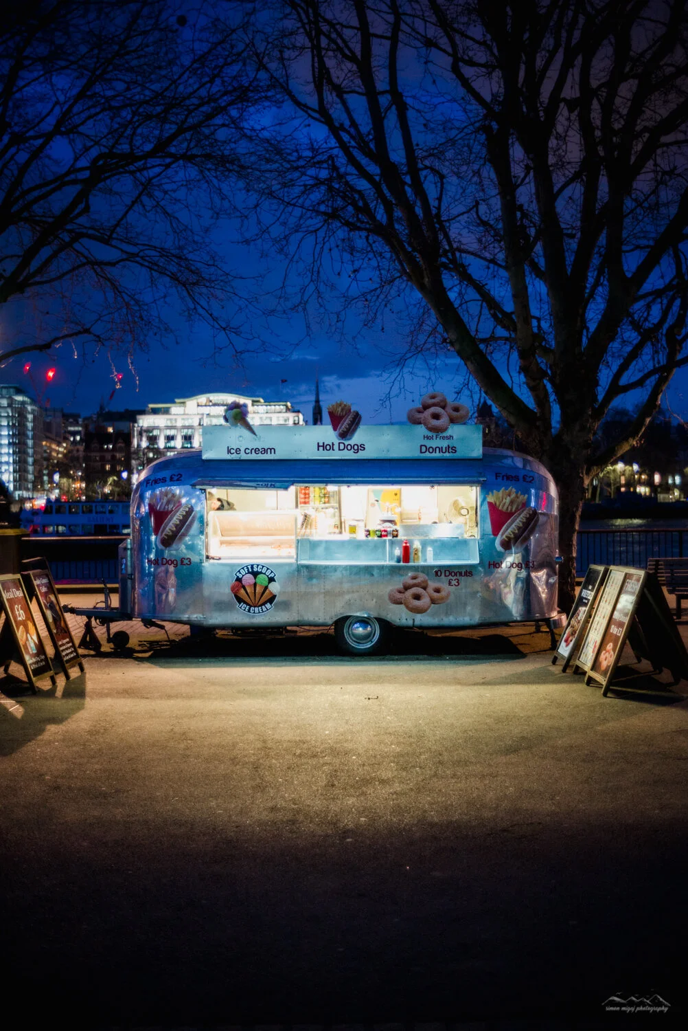 A brightly lit food truck selling ice cream, hot dogs, and donuts, parked under a leafless tree during dusk or night with city buildings and a river in the background.