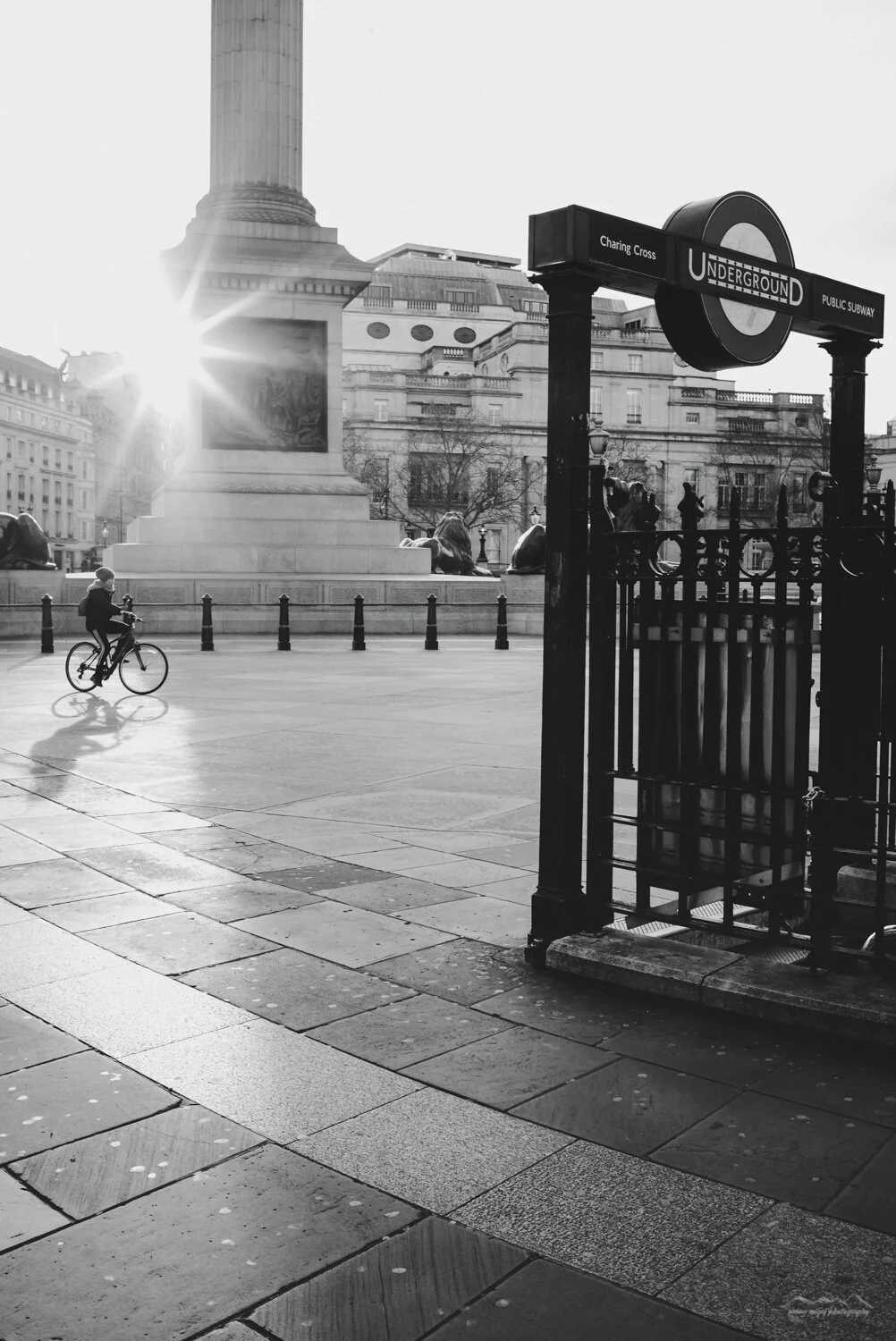 Black and white photo of a city square with a cyclist riding past a monument and a subway entrance marked 'Charing Cross' and 'Underground' in London, England, with buildings and trees in the background and the sun shining brightly.