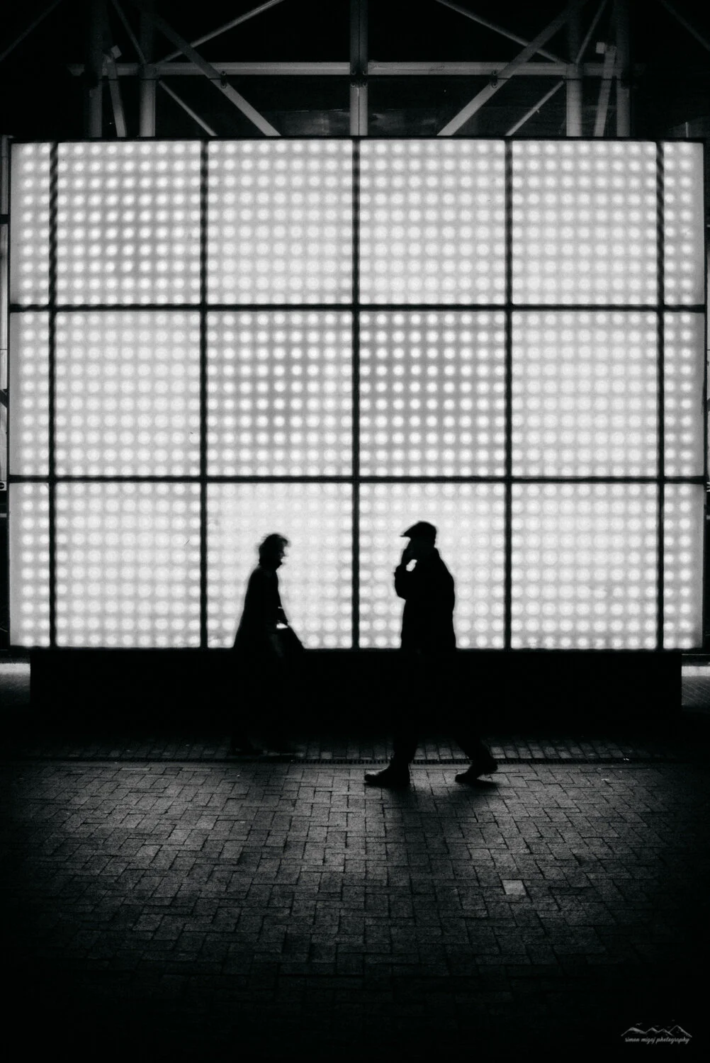 Silhouettes of two people walking and talking in front of a large illuminated grid wall, black and white photo.