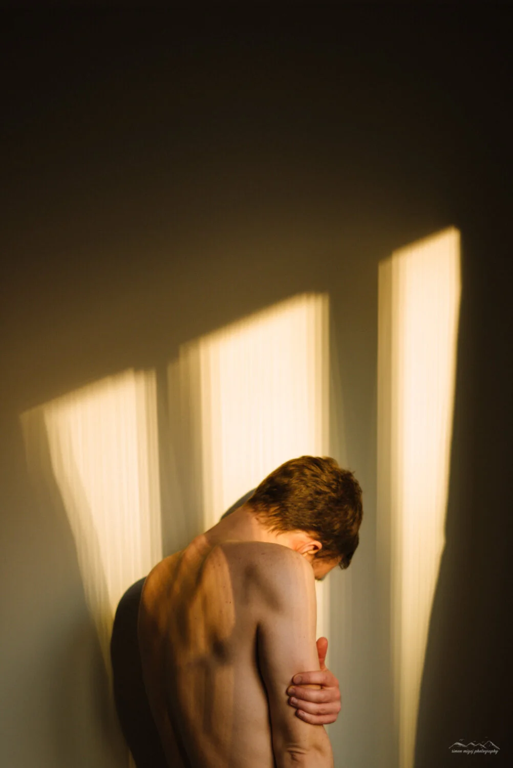 A shirtless man with brown hair is hugging his arm and leaning his head against the wall, illuminated by sunlight coming through blinds, creating vertical patterns of light and shadow.