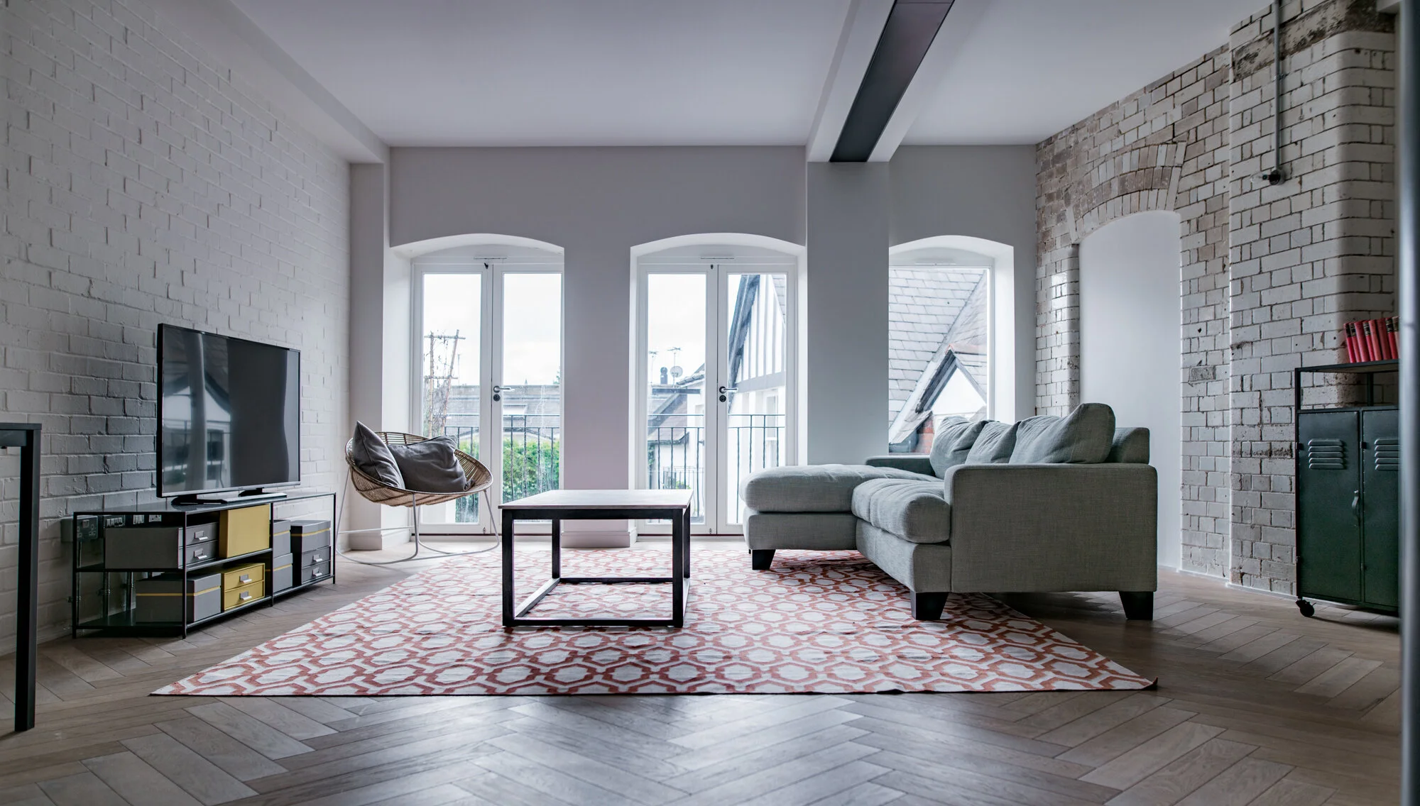 Modern living room with white brick accent wall, large windows, gray sofa, patterned area rug, and a flat-screen TV.
