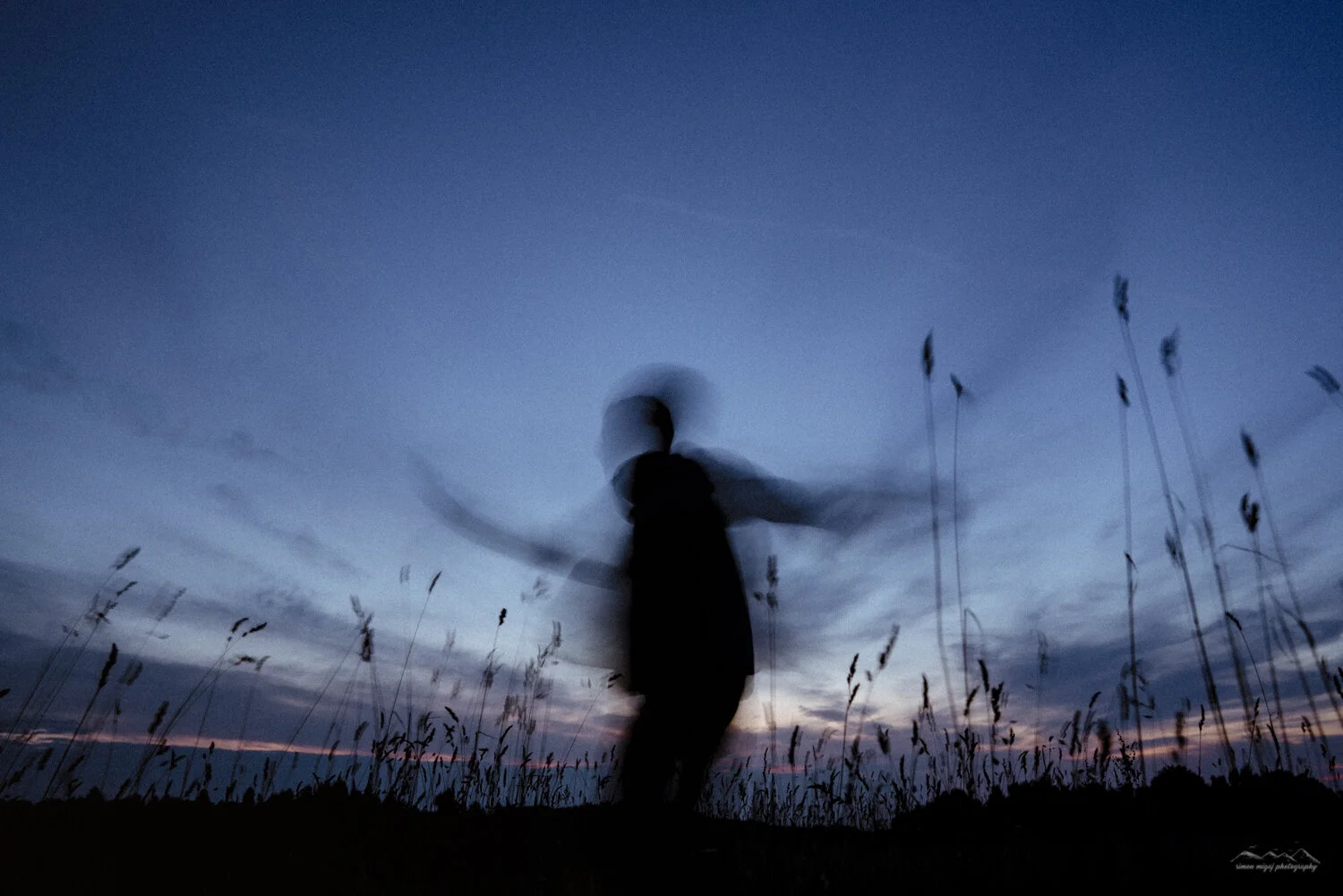 A blurred silhouette of a person standing in a field of tall grasses during dusk or dawn, with a dark sky and some clouds in the background.