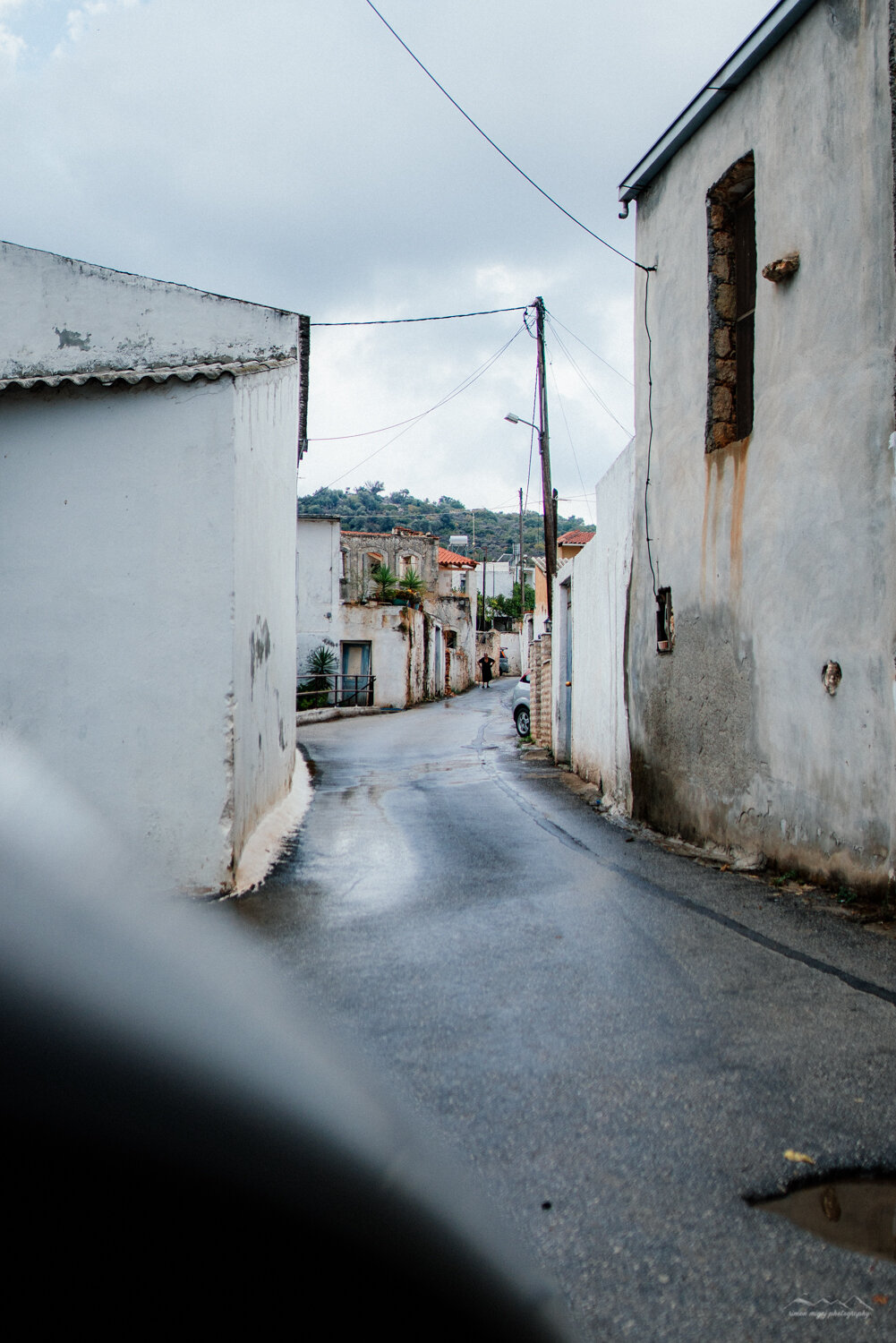 A narrow paved street in a small town, with white and beige buildings on either side, some with weathered walls and small windows, under a cloudy sky, with a person walking in the distance.