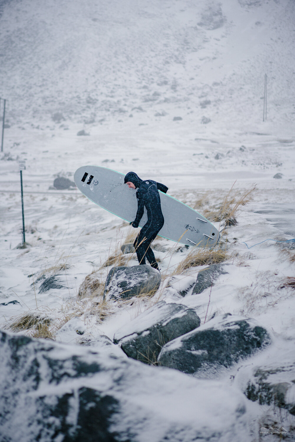 A person in a black wetsuit carrying a surfboard on a snowy, rocky landscape with snow falling.