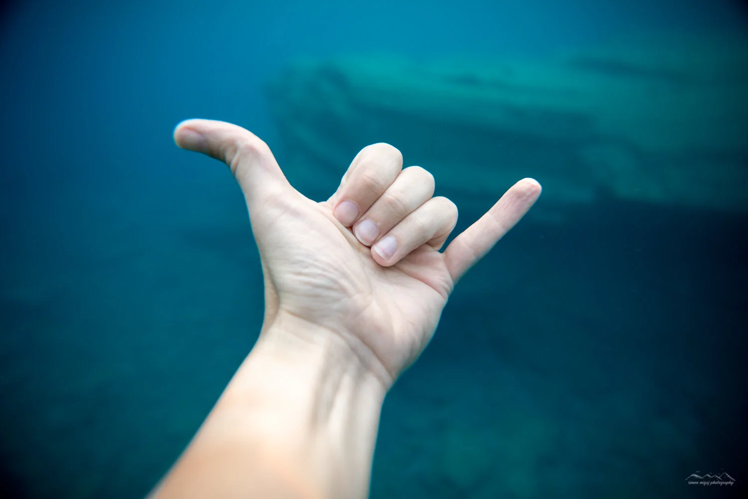 A human hand underwater making the 'shaka' gesture with fingers curled and thumb and pinky finger extended.
