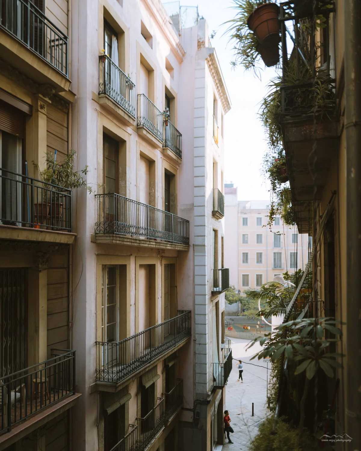 View of beige residential buildings with black balconies and potted plants, taken from a window overlooking a courtyard with a woman walking and a child playing.
