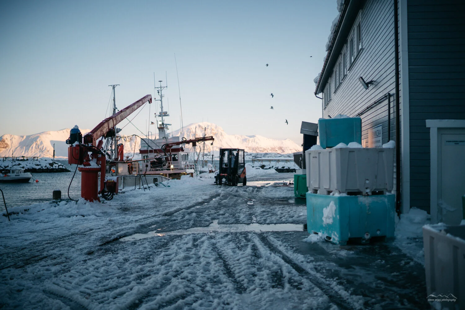Snowy dock area with boats, a forklift, and storage containers, with mountains in the background and a clear sky.