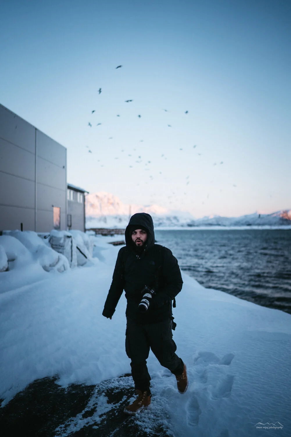 A person dressed in black cold-weather clothing, holding a camera with a long lens, standing on snowy ground near a body of water, with a building on the left and a flock of birds flying overhead against a mountain backdrop.