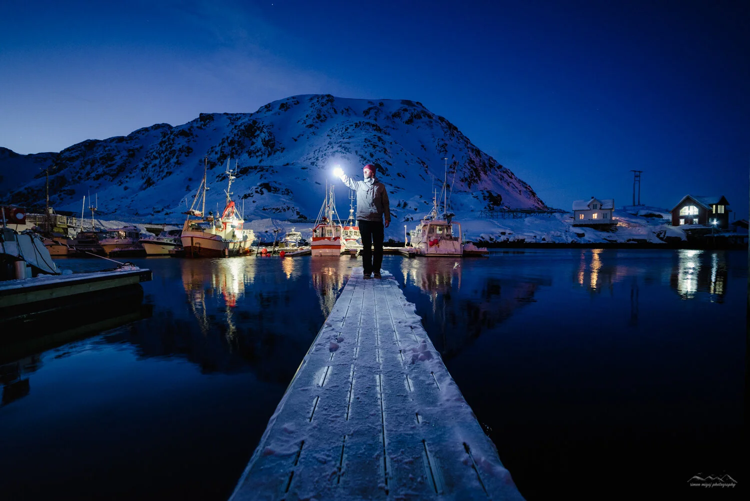 Person standing on a snow-covered dock at night, holding a flashlight with boats and snow-covered mountains in the background.