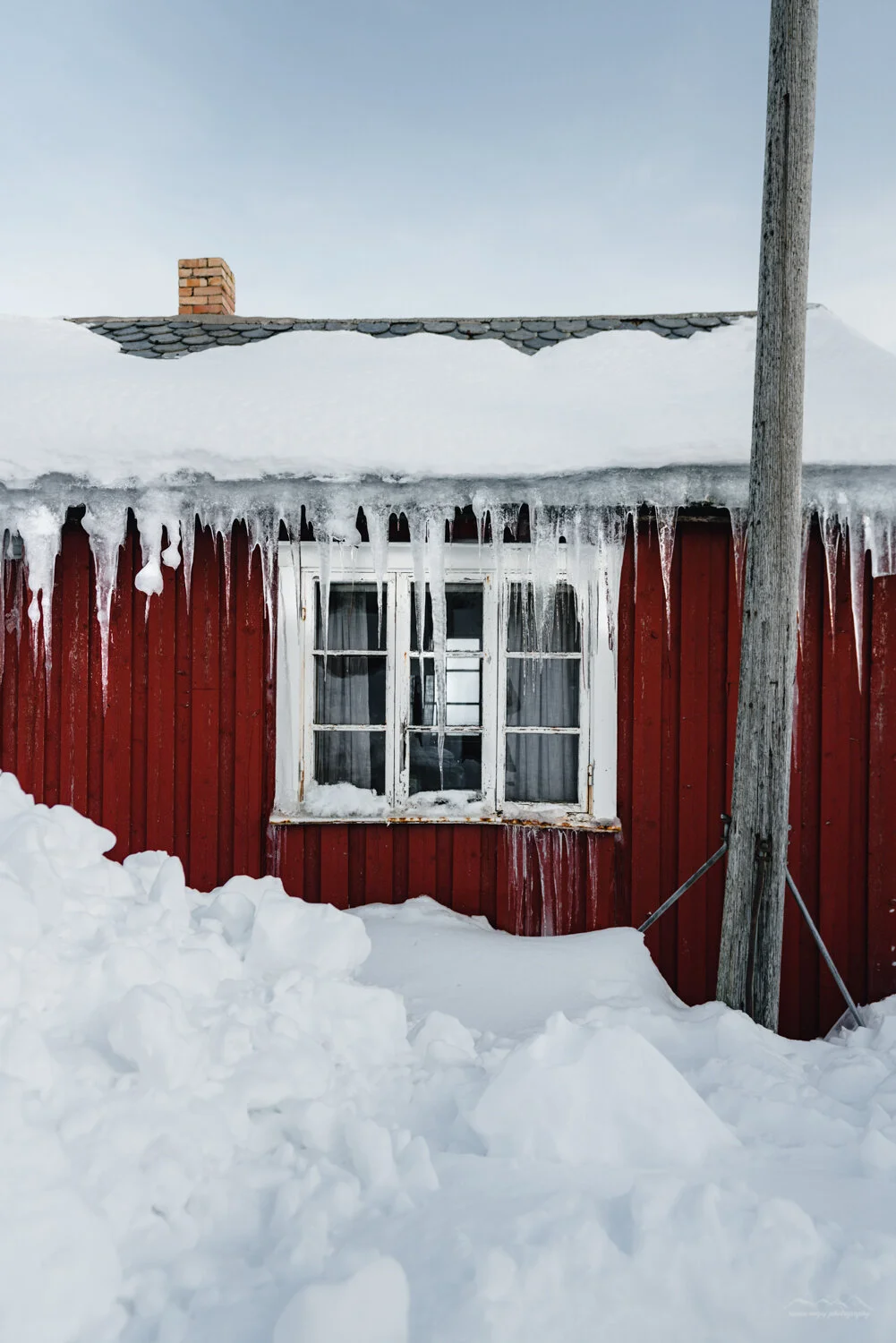 A red wooden house covered in snow and ice with icicles hanging from the roof, a window, and a utility pole in front.