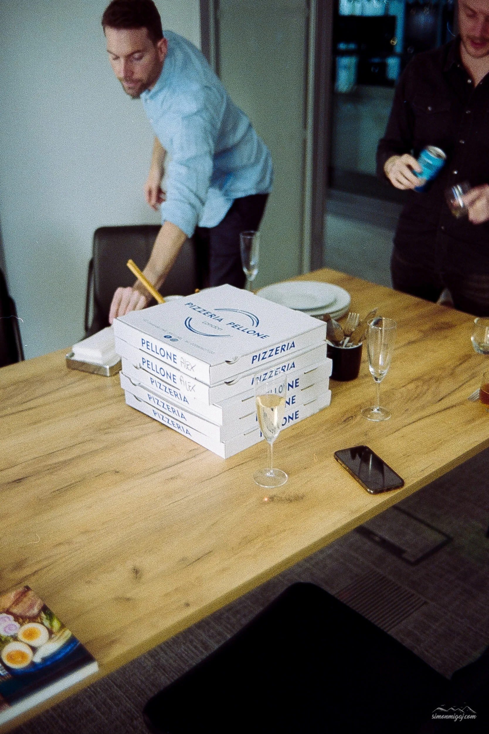 A group of people gathered around a wooden table with pizza boxes labeled "Pizzeria Pellone" and glasses of champagne.