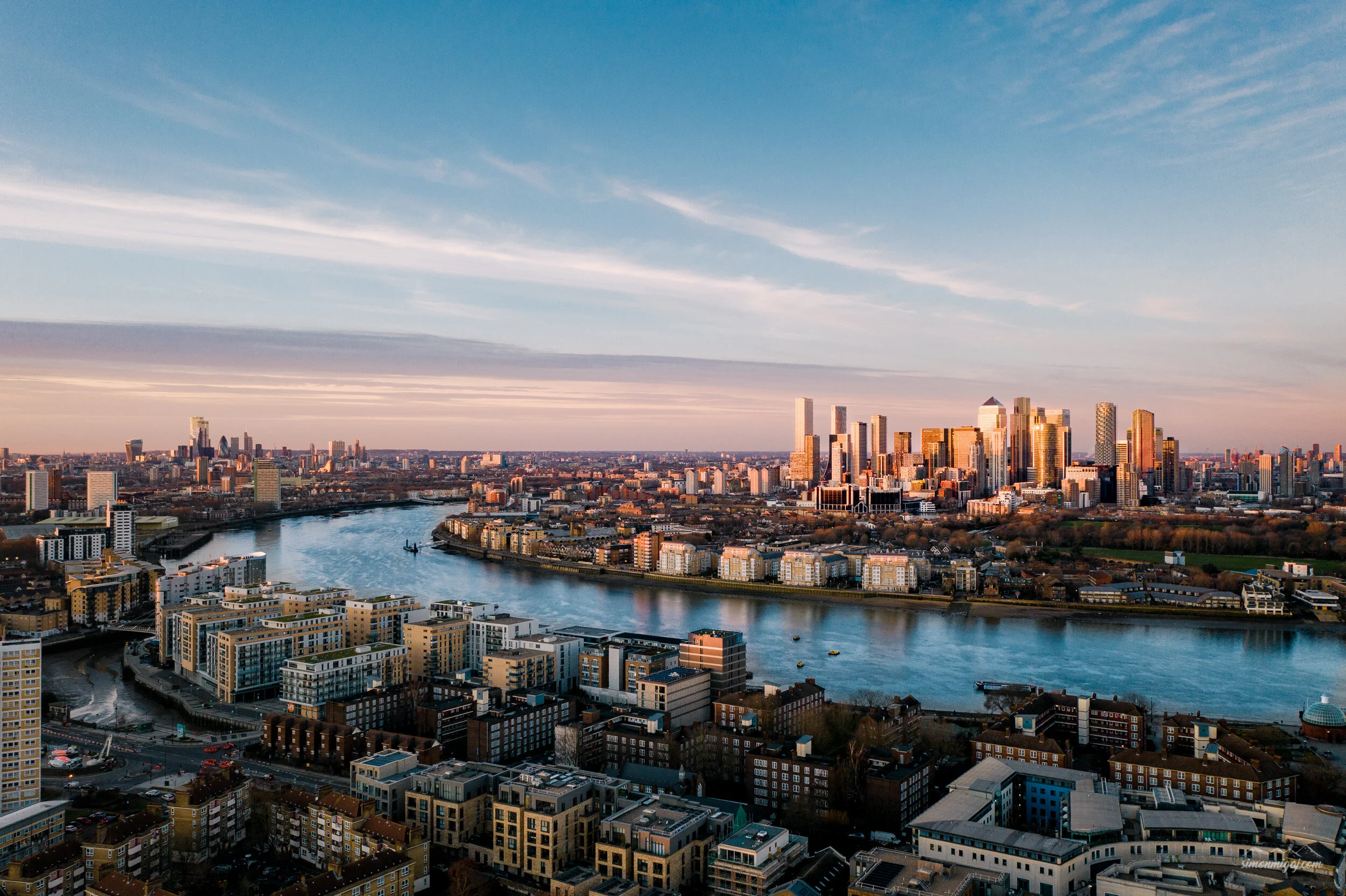 A panoramic view of a city skyline with tall skyscrapers, a river flowing through the city, and modern residential buildings in the foreground. The sky is partly cloudy with a gradient of blue and pink hues from the setting or rising sun.