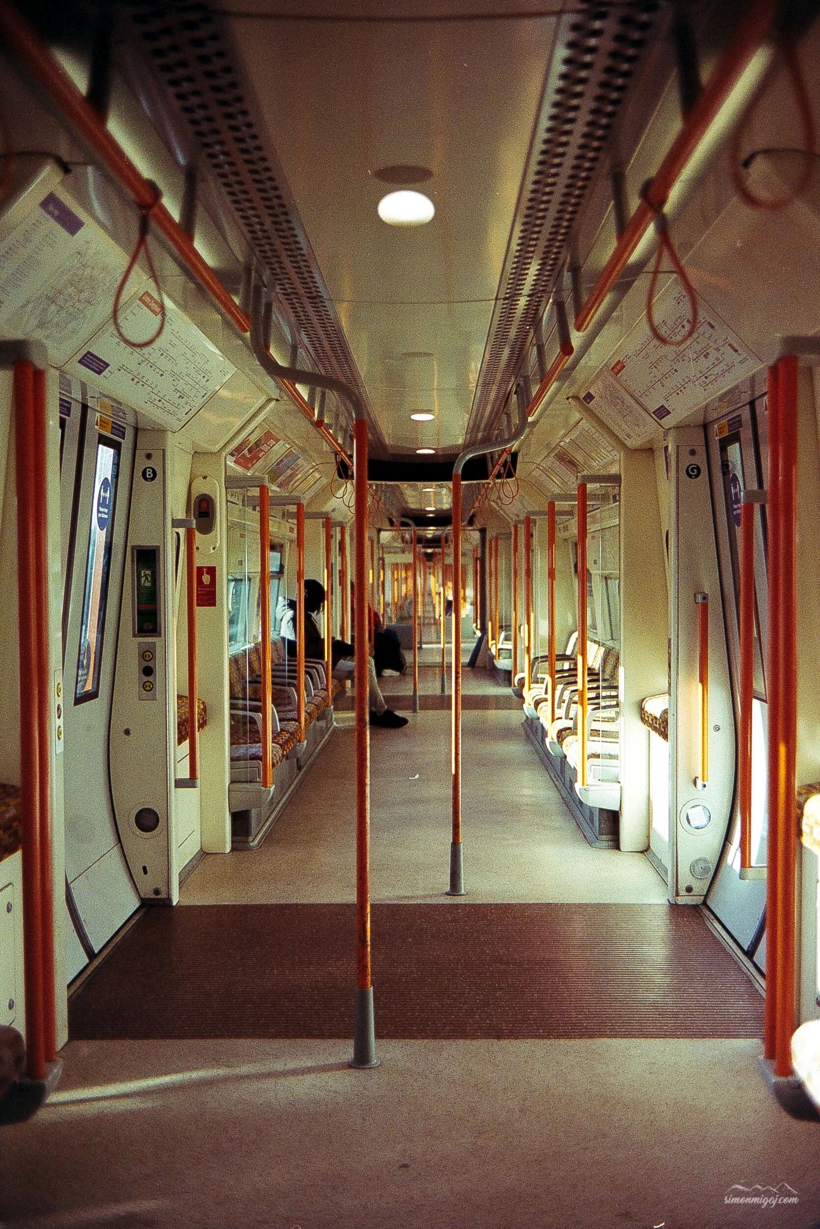 Interior of a public transit bus or train with orange poles and seats, some passengers seated, and information panels on the walls.