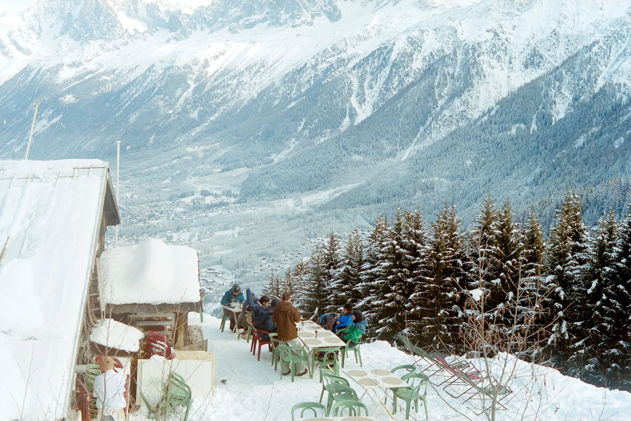 People gathered outdoors on a snowy mountain slope, sitting at tables and socializing, with snow-covered trees and mountain peaks in the background.