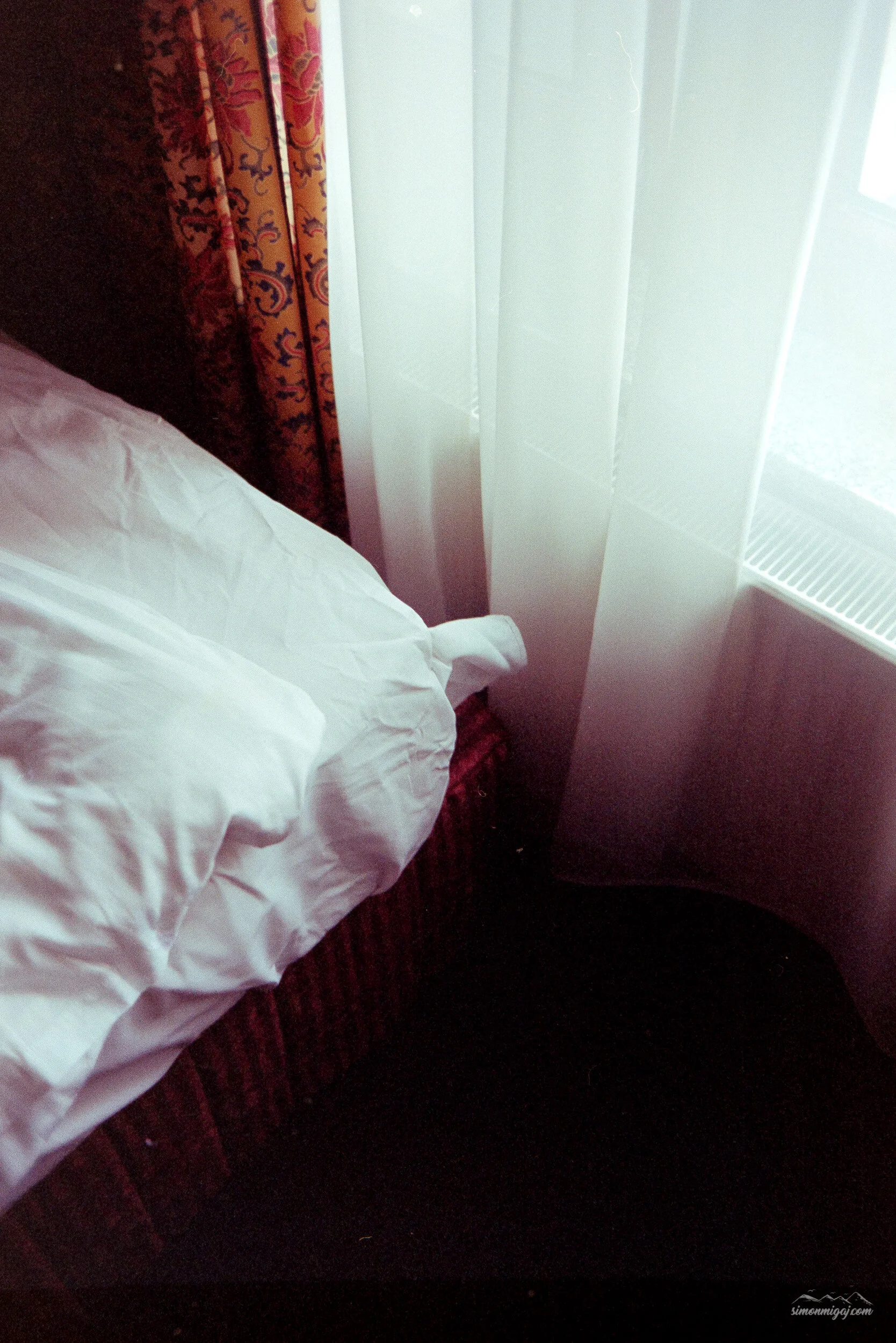 Part of a bed with white sheets, a red and black patterned carpet, a window with patterned curtains, and a white radiator below the window.