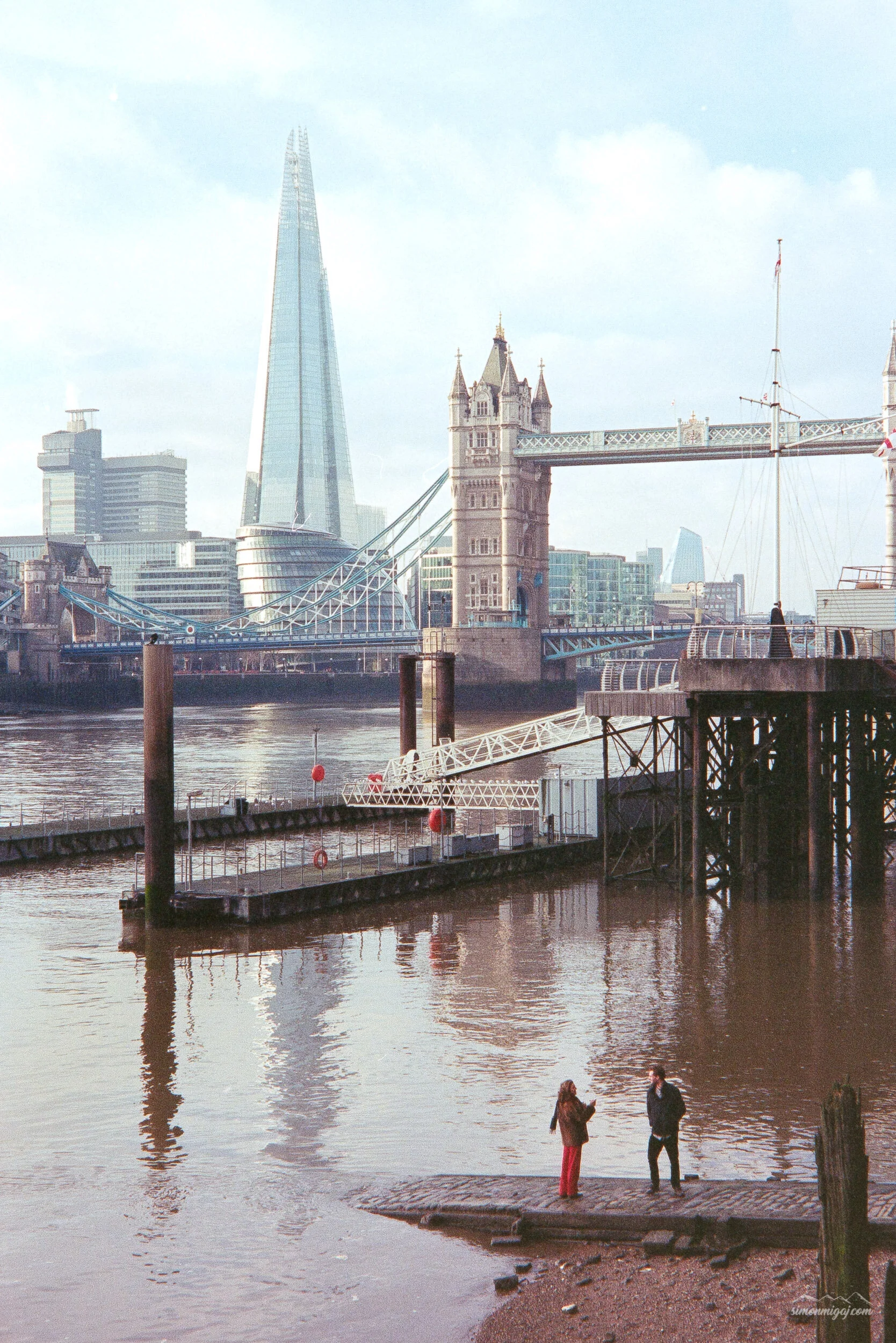 People walking along the riverbank near Tower Bridge in London with the Shard skyscraper in the background on a cloudy day.