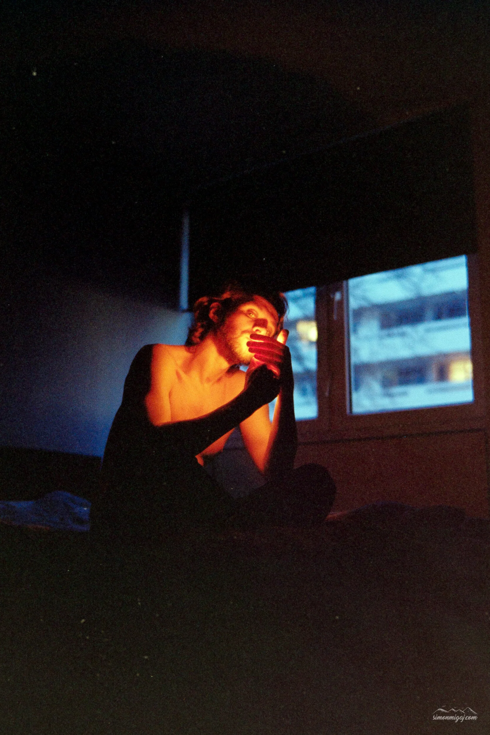A person with short wavy hair and a beard sitting on the floor in a dimly lit room, with their hand covering part of their face, illuminated by a soft, warm light. There is a window in the background showing an outdoor scene with a building.