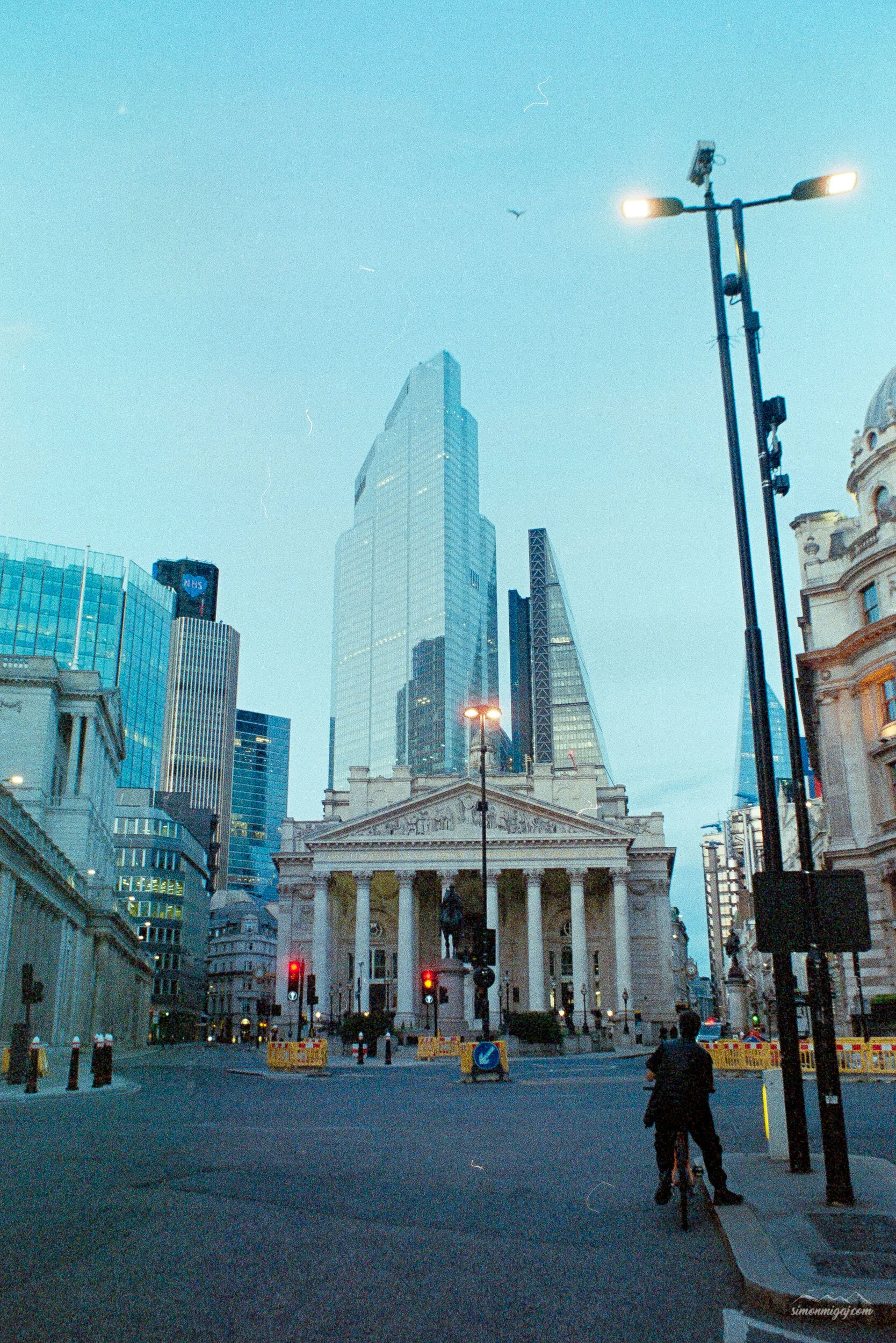 City street scene with tall modern skyscrapers and a historic building with columns in the foreground, streetlights, and a man on a bicycle at the corner during dusk.