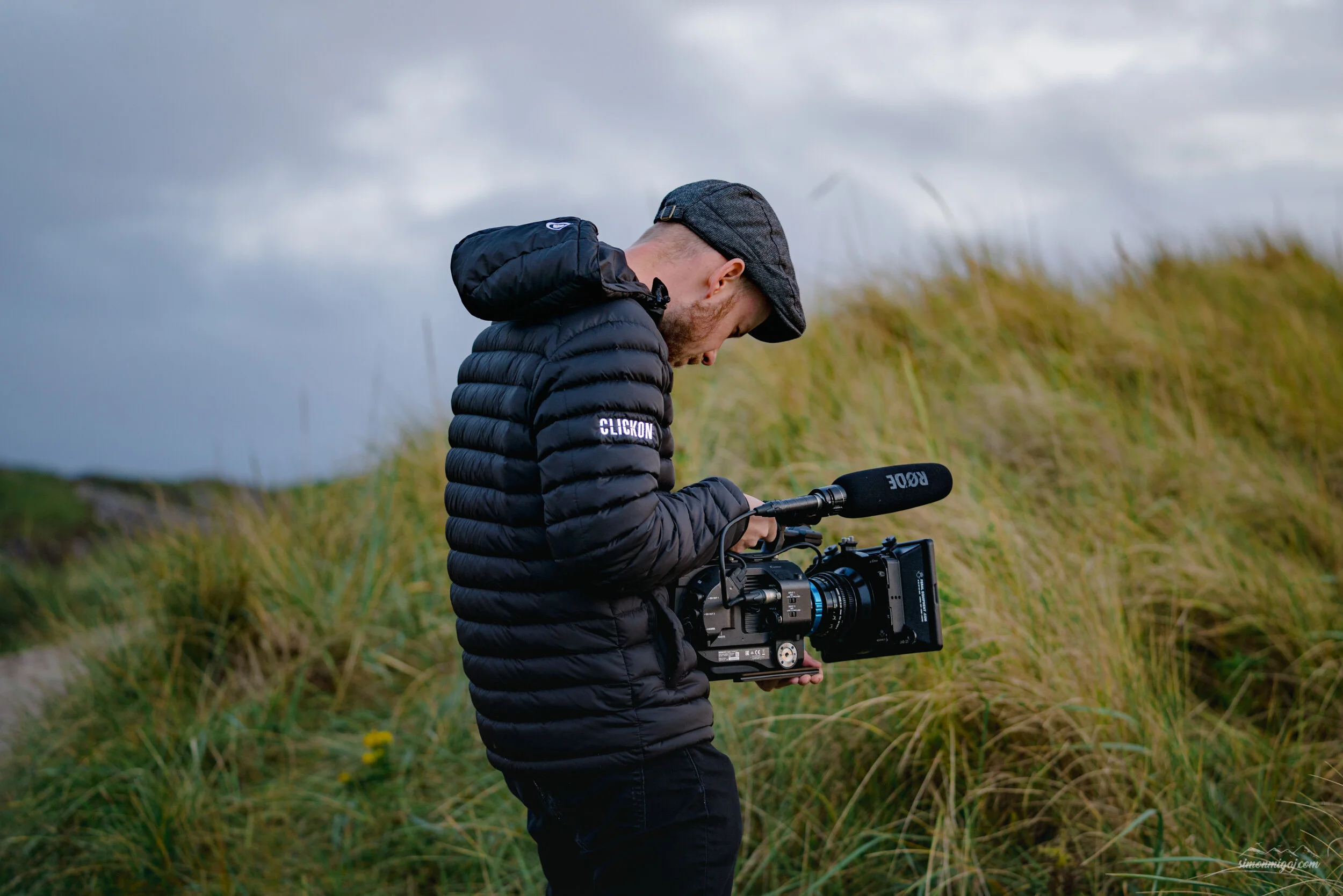 A man wearing a dark jacket with 'CLICK ON' written on the sleeve, a gray cap, and black pants, is holding a professional video camera outdoors on a grassy field with cloudy sky in the background.