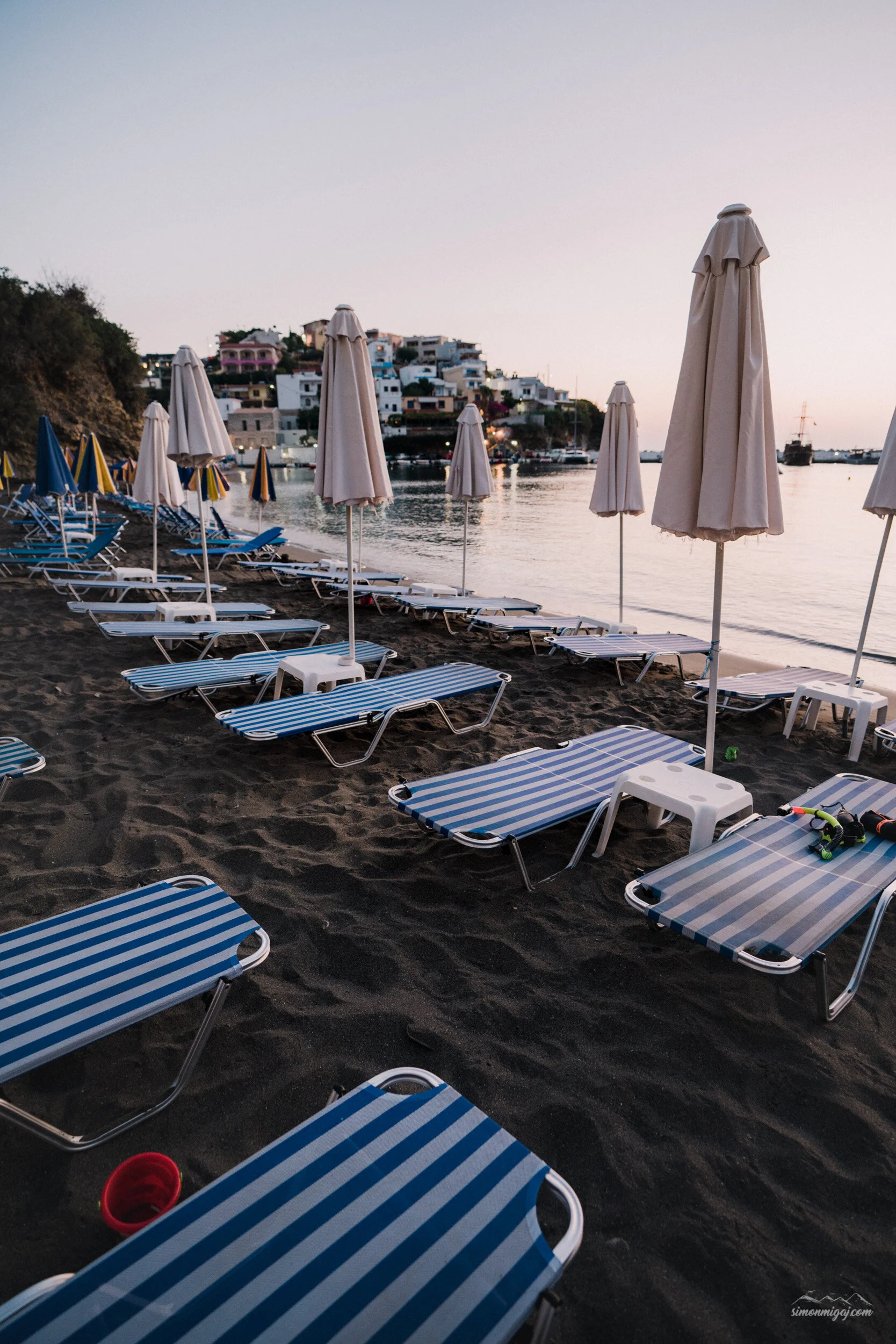 Empty beach chairs with blue and white stripes and beige umbrellas lined along a shore at sunset, with a hillside town of white and pastel buildings in the background and calm water reflecting the sky.