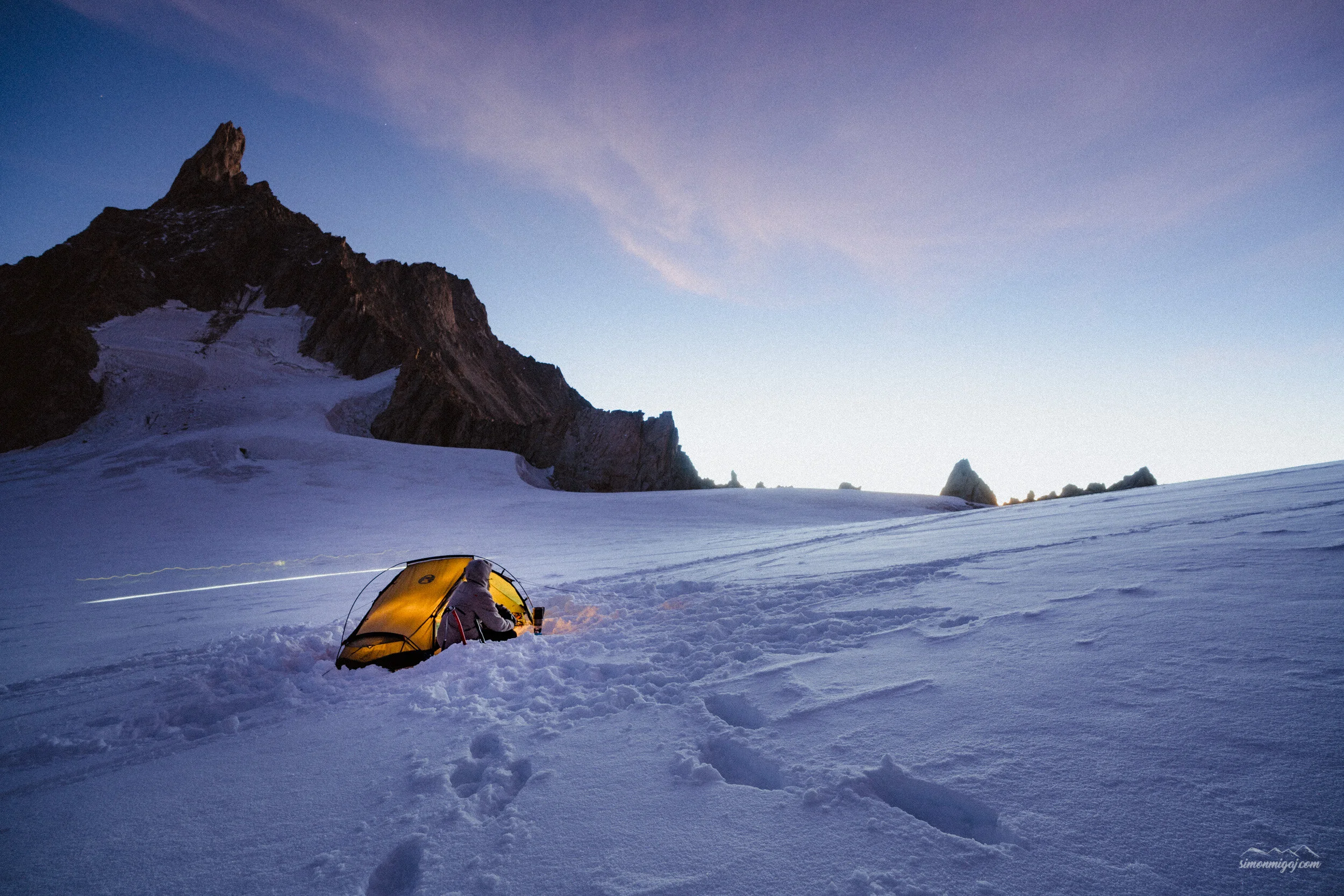 A person camping on a snowy mountain at sunset with a yellow tent, snow-covered ground, and jagged mountain peaks in the background.