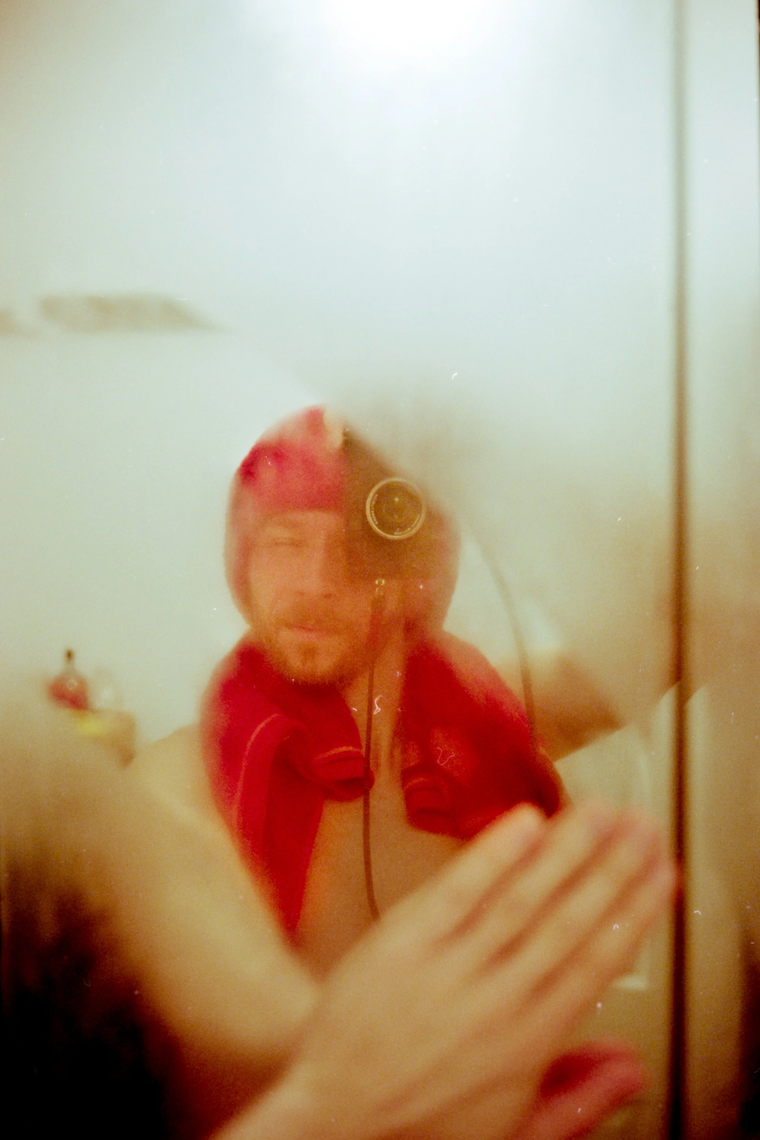 A person taking a mirror selfie with a phone inside a bathroom, wearing a red hoodie. The person's face is partially visible and blurred.