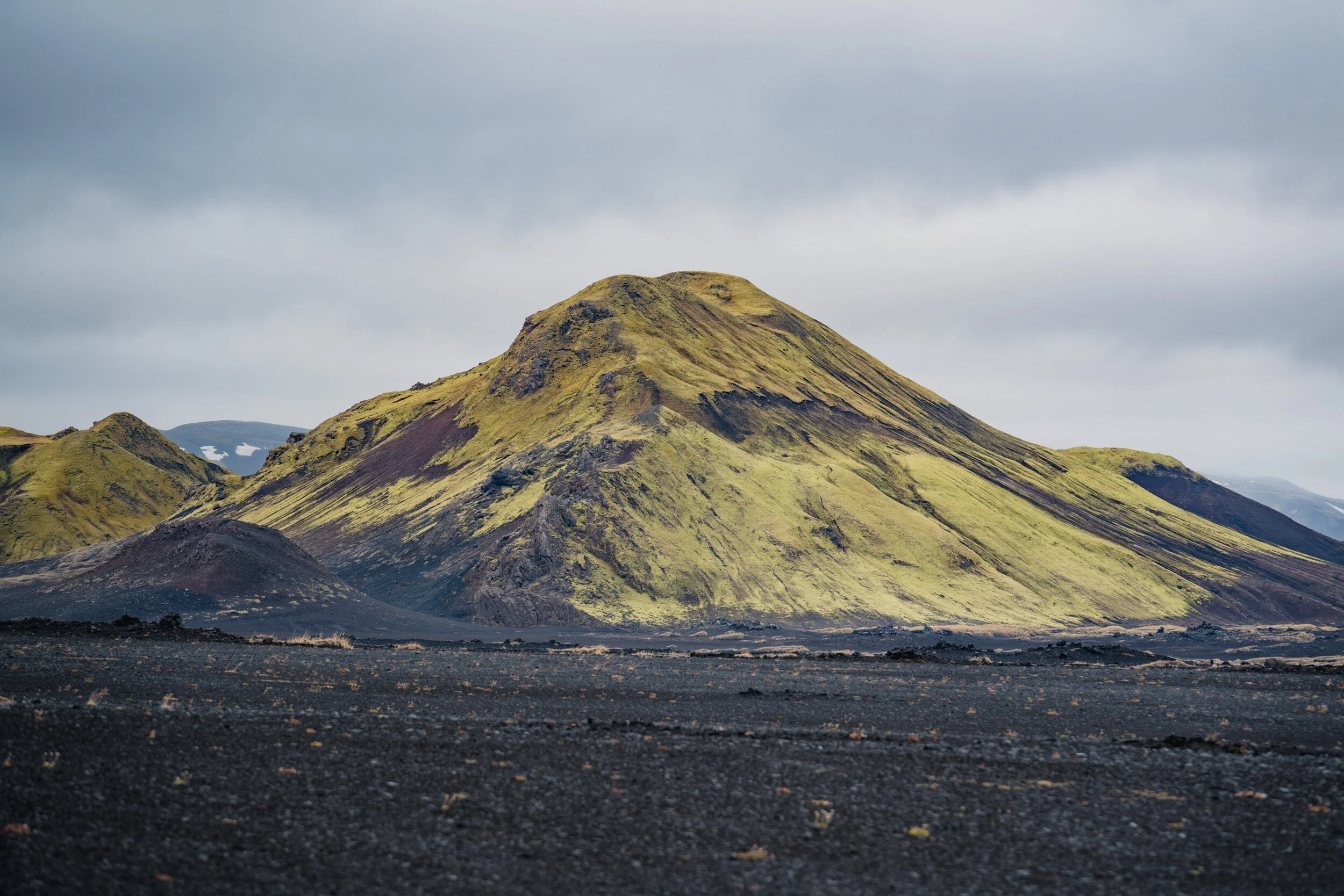 A moss-covered volcanic mountain with a dark base, set against a cloudy sky.